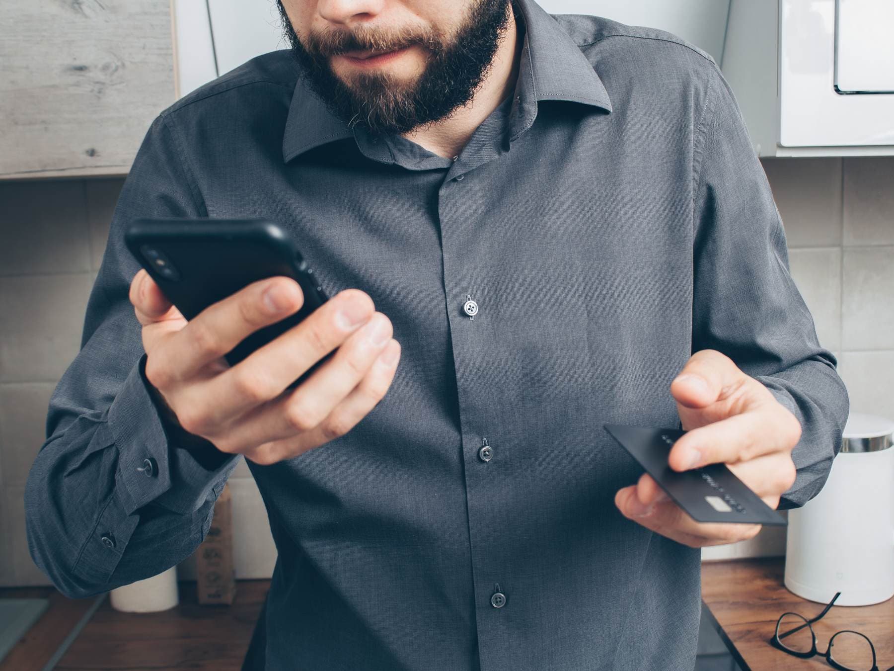 Uomo con barba e camicia grigia che tiene uno smartphone in una mano e una carta di pagamento nell'altra, in una cucina