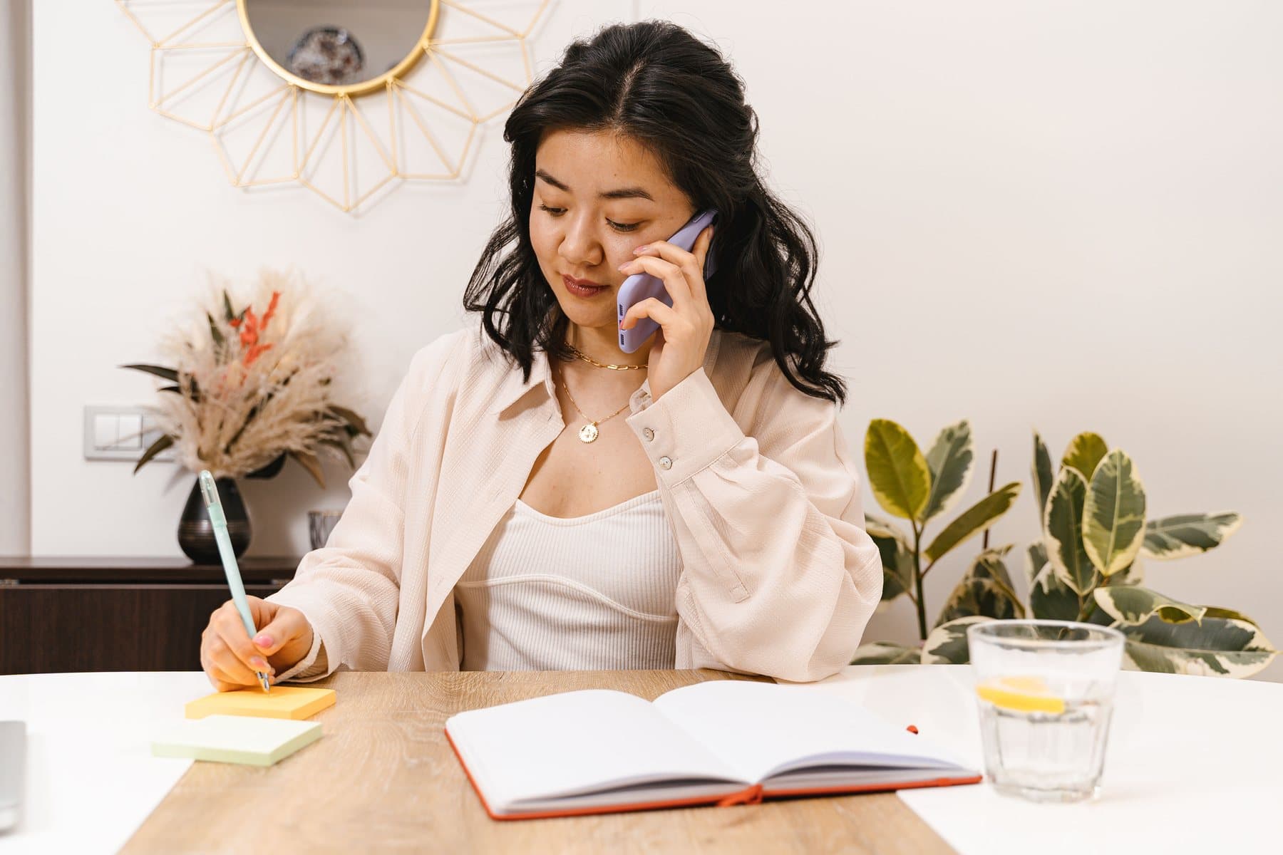 A woman sitting at a table takes notes on a sticky note while talking on the phone