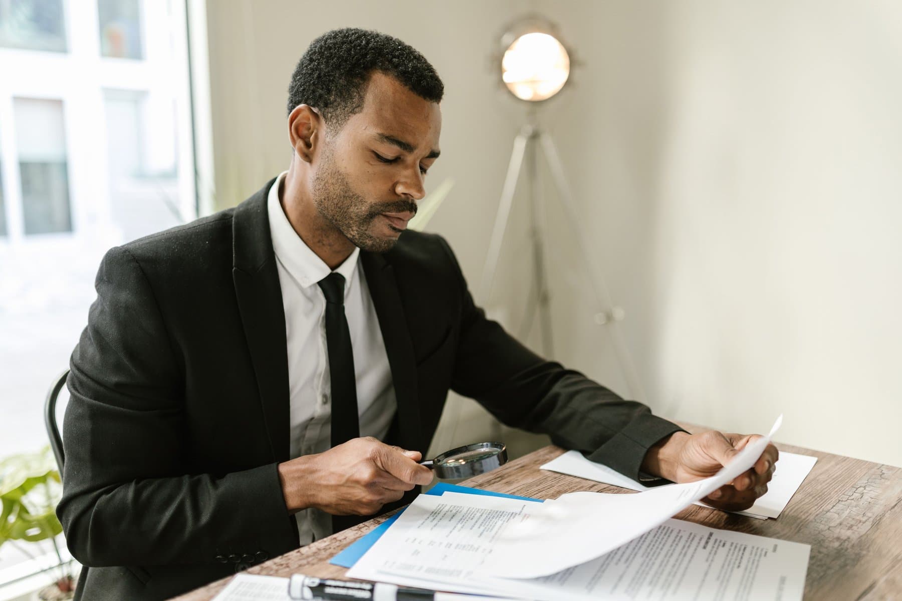 A man in a suit and tie examines a printed document with a magnifying glass, seated at a desk