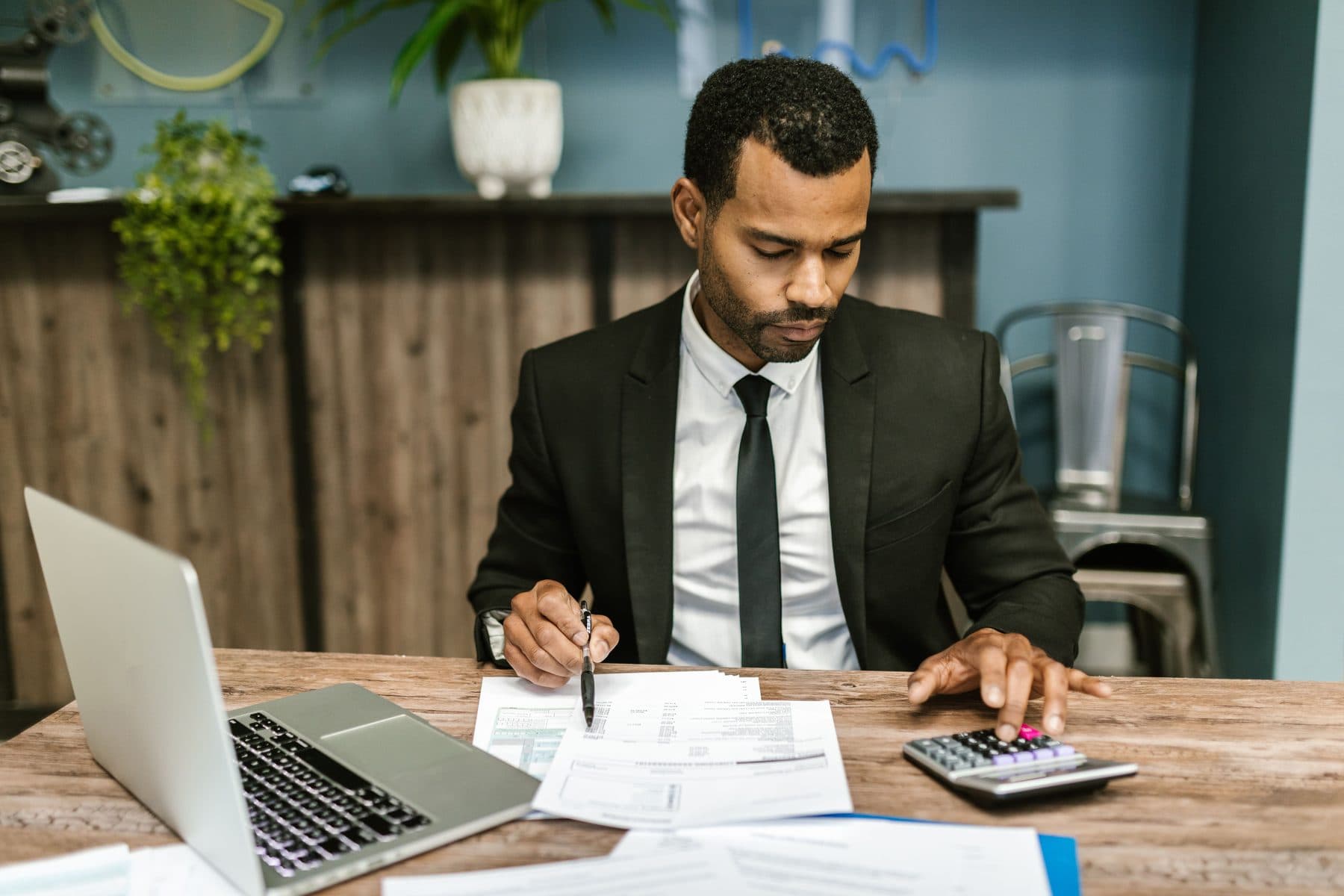 A man in a suit checks invoices with a calculator, in front of an open laptop on the desk