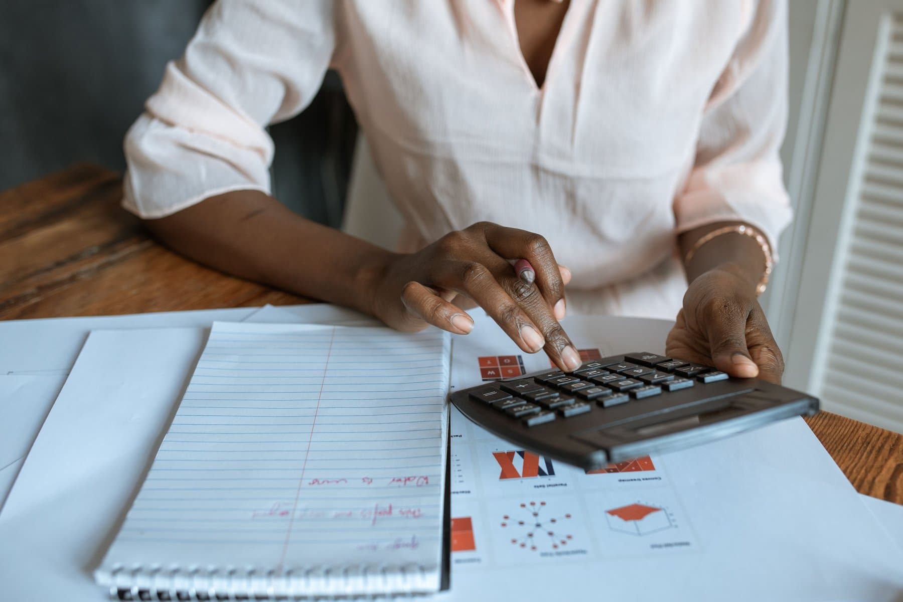 A person uses a calculator at a desk, with a notepad and printed spreadsheets next to it