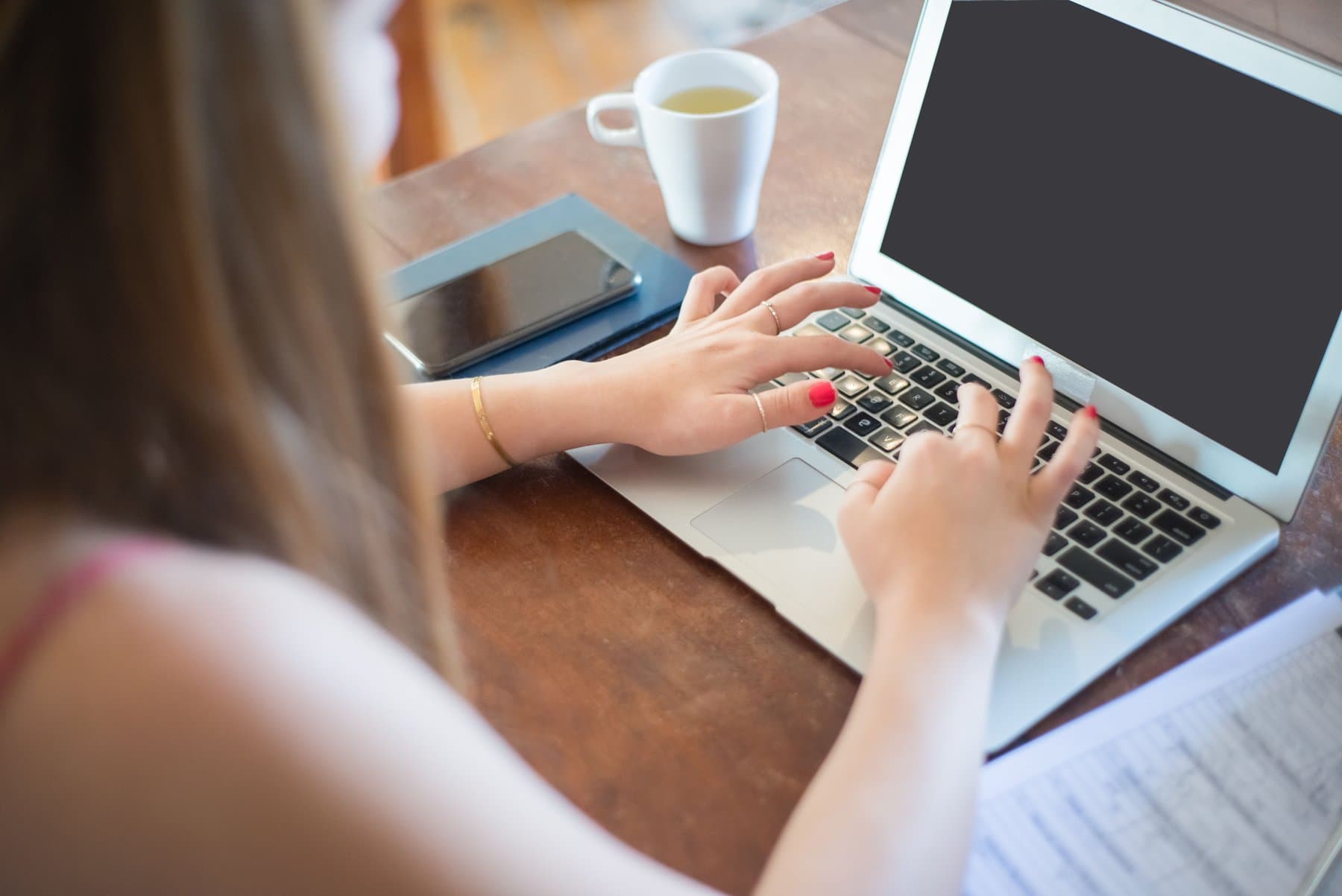 Hands typing on a laptop keyboard, with a smartphone resting on the desk next to a cup of tea