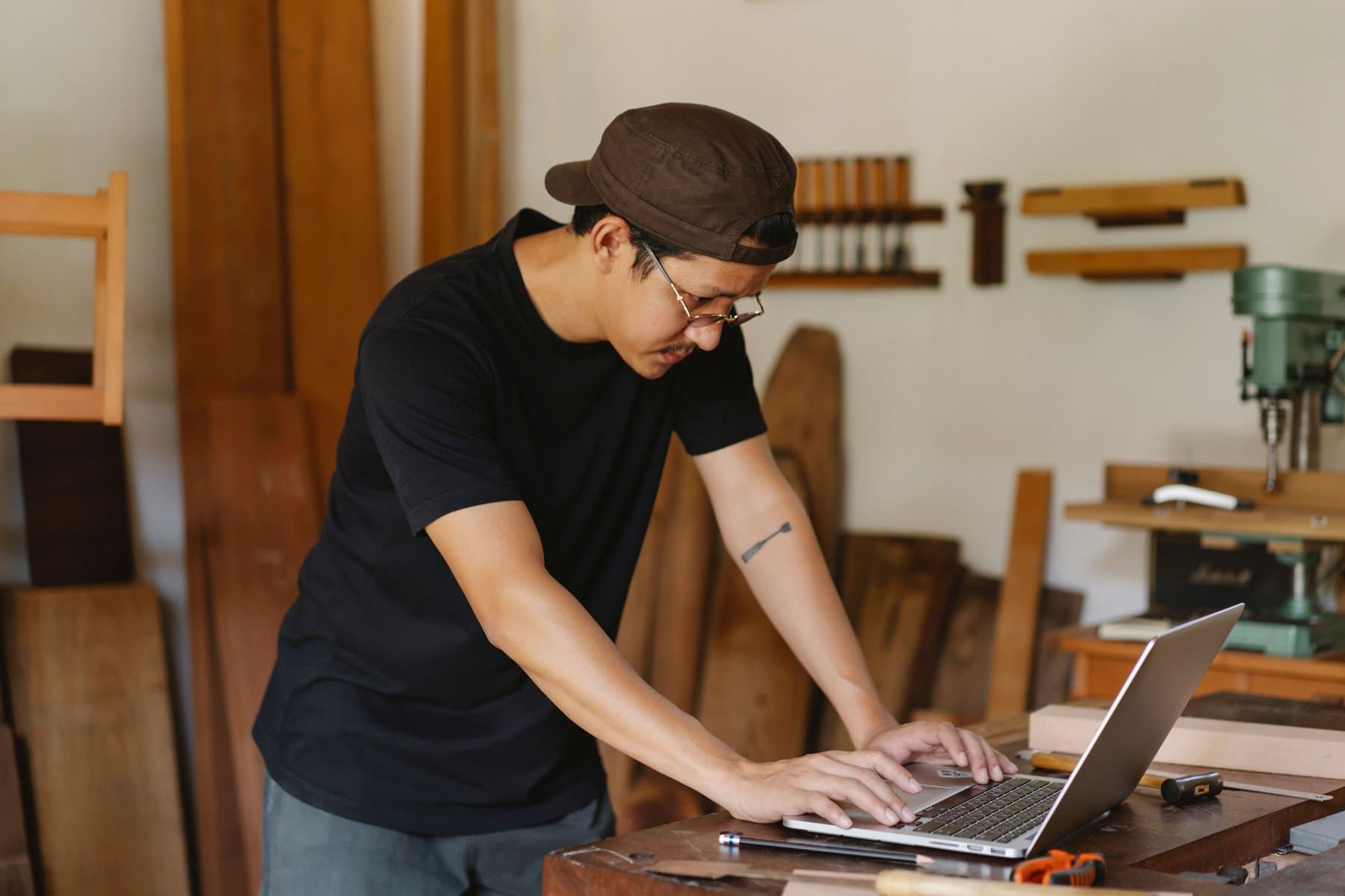 A craftsman in a woodworking shop types on a laptop resting on his workbench, surrounded by tools and wood planks