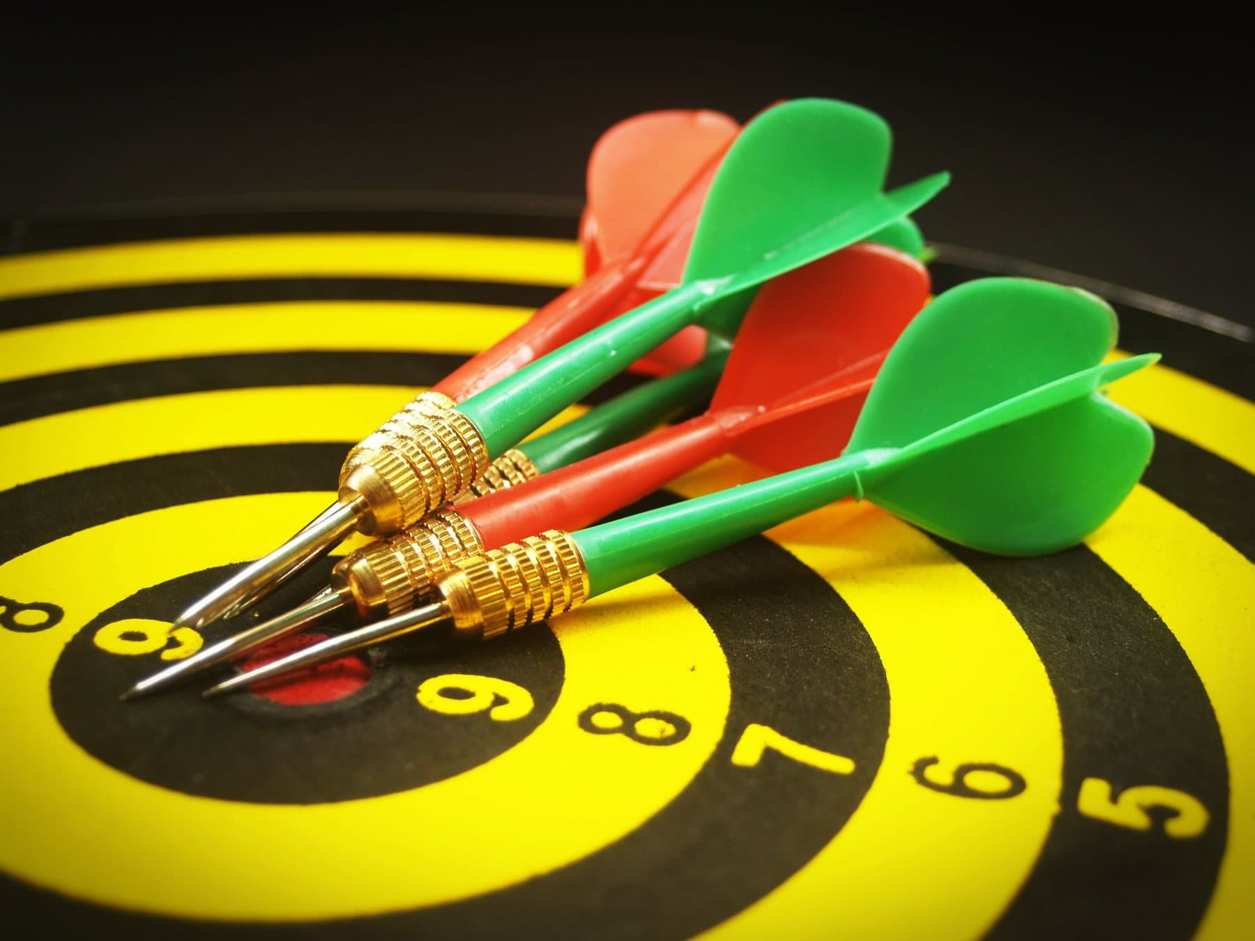 Three darts, two green and one red, stuck in the centre of a yellow and black dartboard