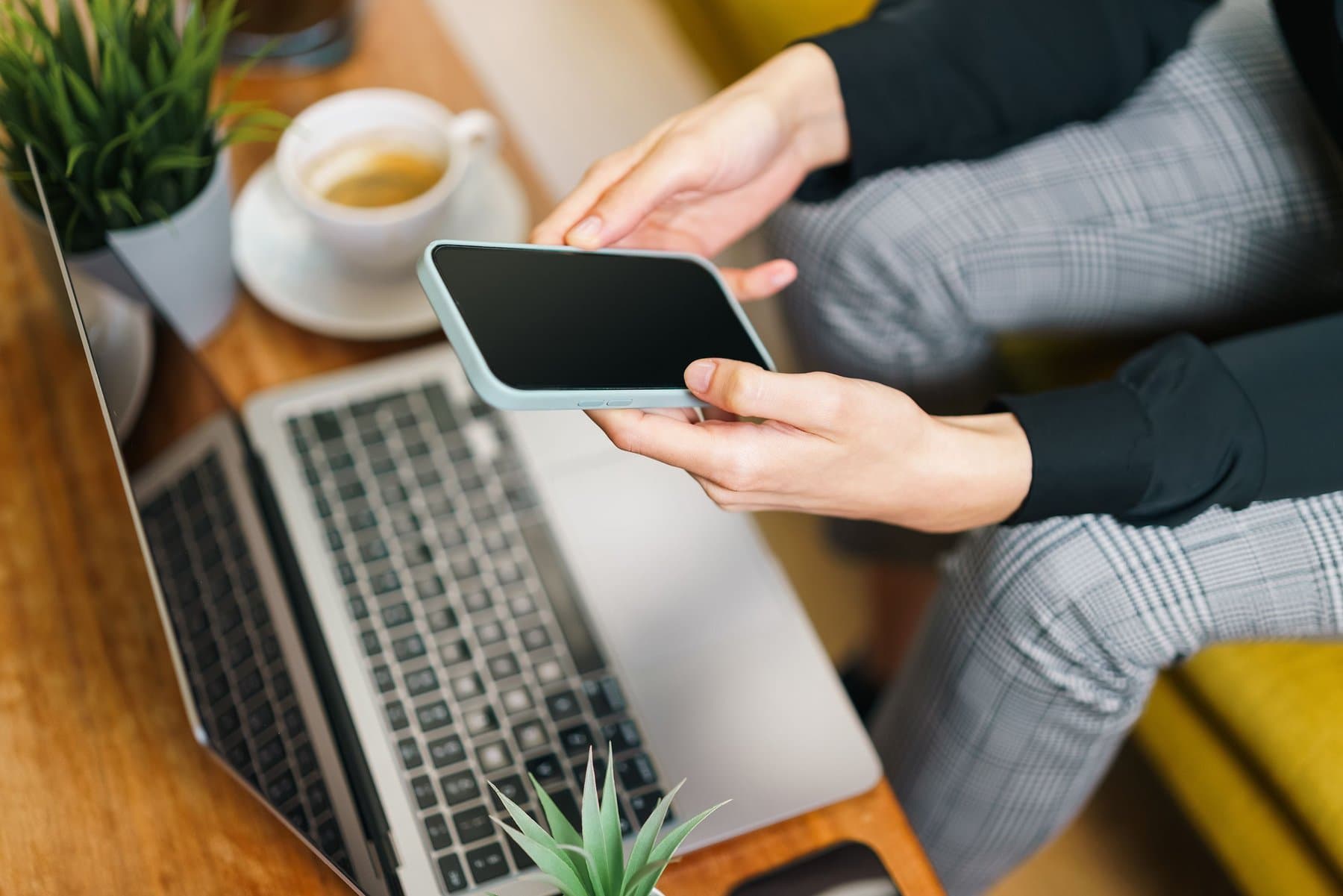 A person holds a smartphone with the screen off, above an open laptop on a desk with a cup of coffee and a small plant