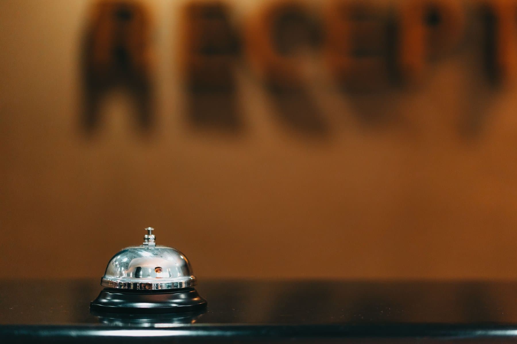 A silver reception call bell in close-up on a dark counter, with a blurred "Reception" sign behind