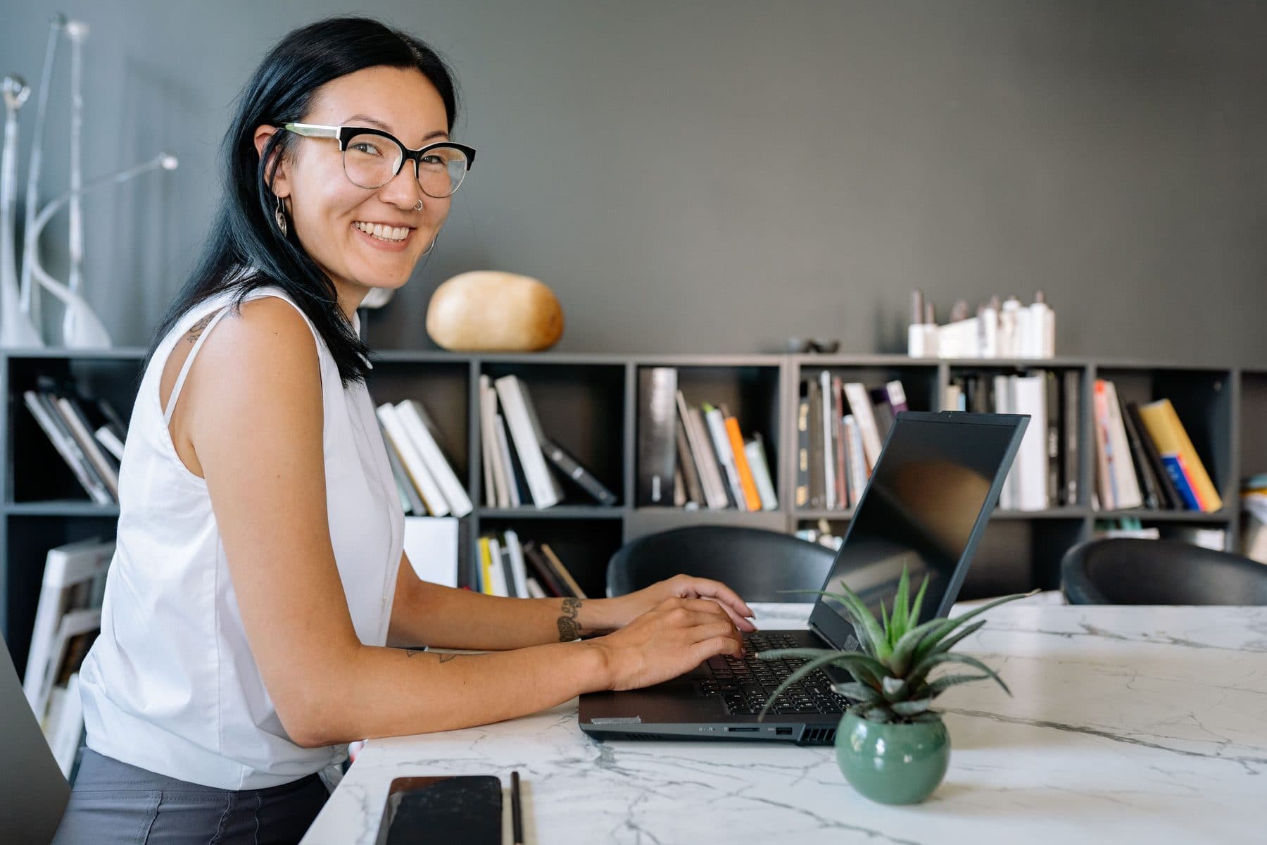 A woman smiles while working on a laptop in a modern office with a bookshelf in the background