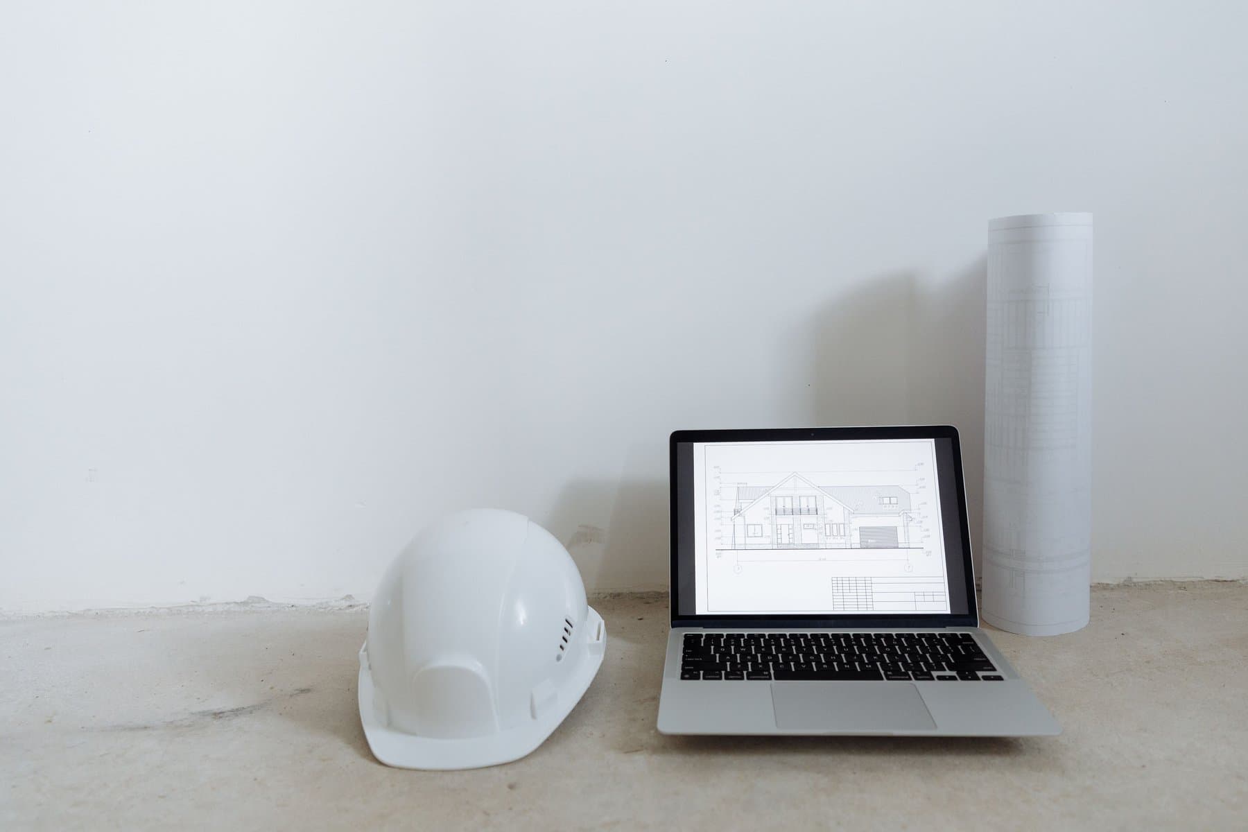 A white construction hard hat next to an open laptop showing the technical drawing of a house, with a rolled-up blueprint in the background, on a bare concrete floor