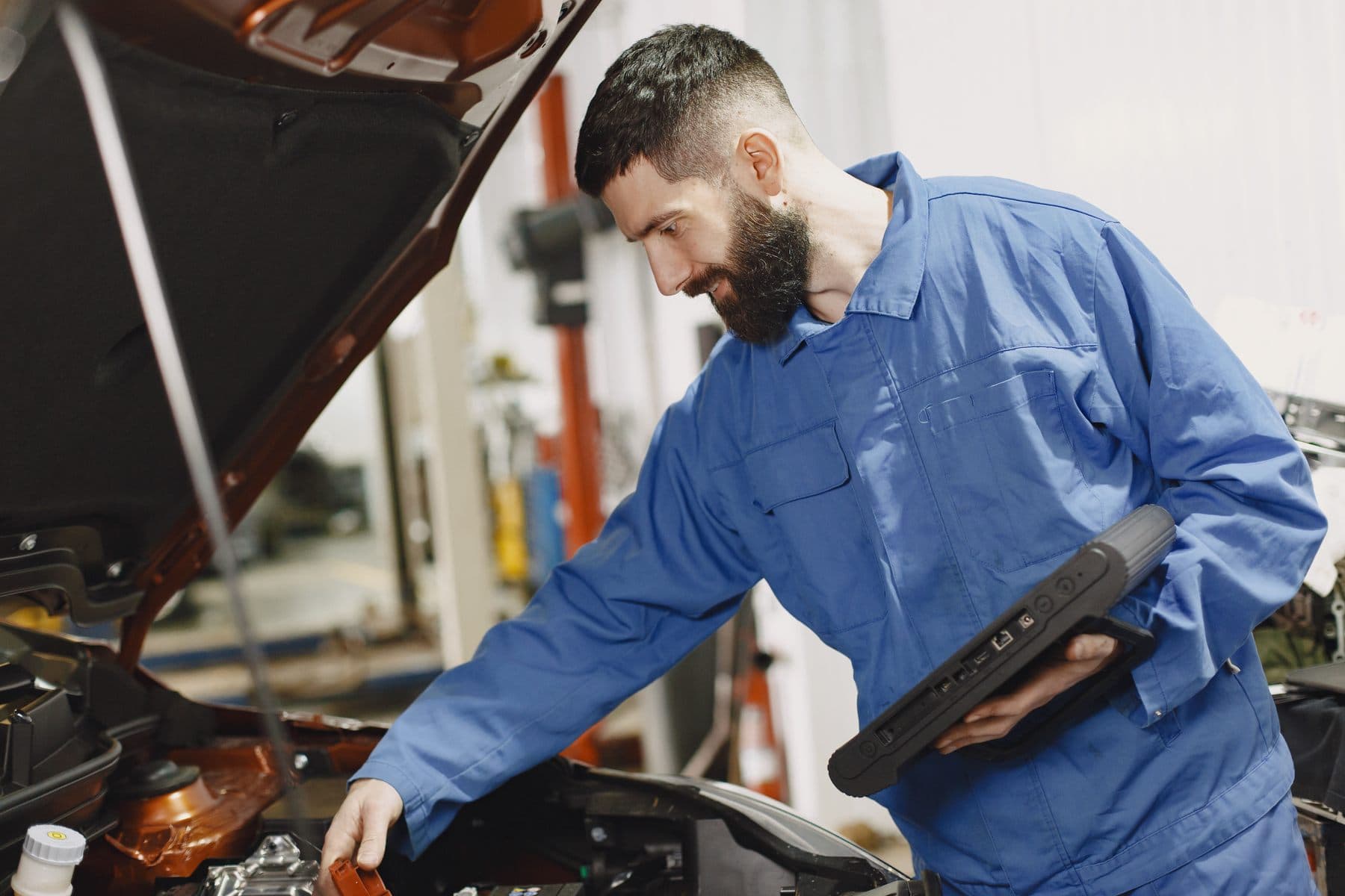 A bearded mechanic in blue overalls leans over an open car engine bay while holding a rugged diagnostic scanner