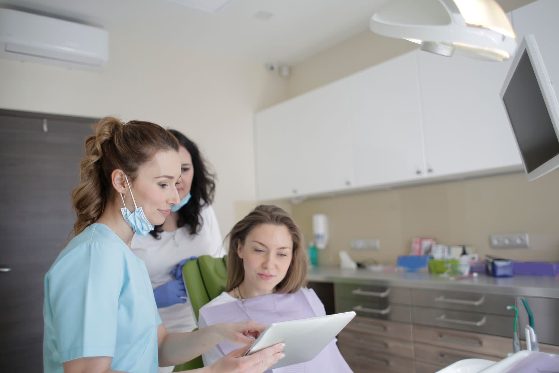 A dentist shows a tablet to a patient seated in the dental chair, with an assistant standing beside her, in a bright modern practice