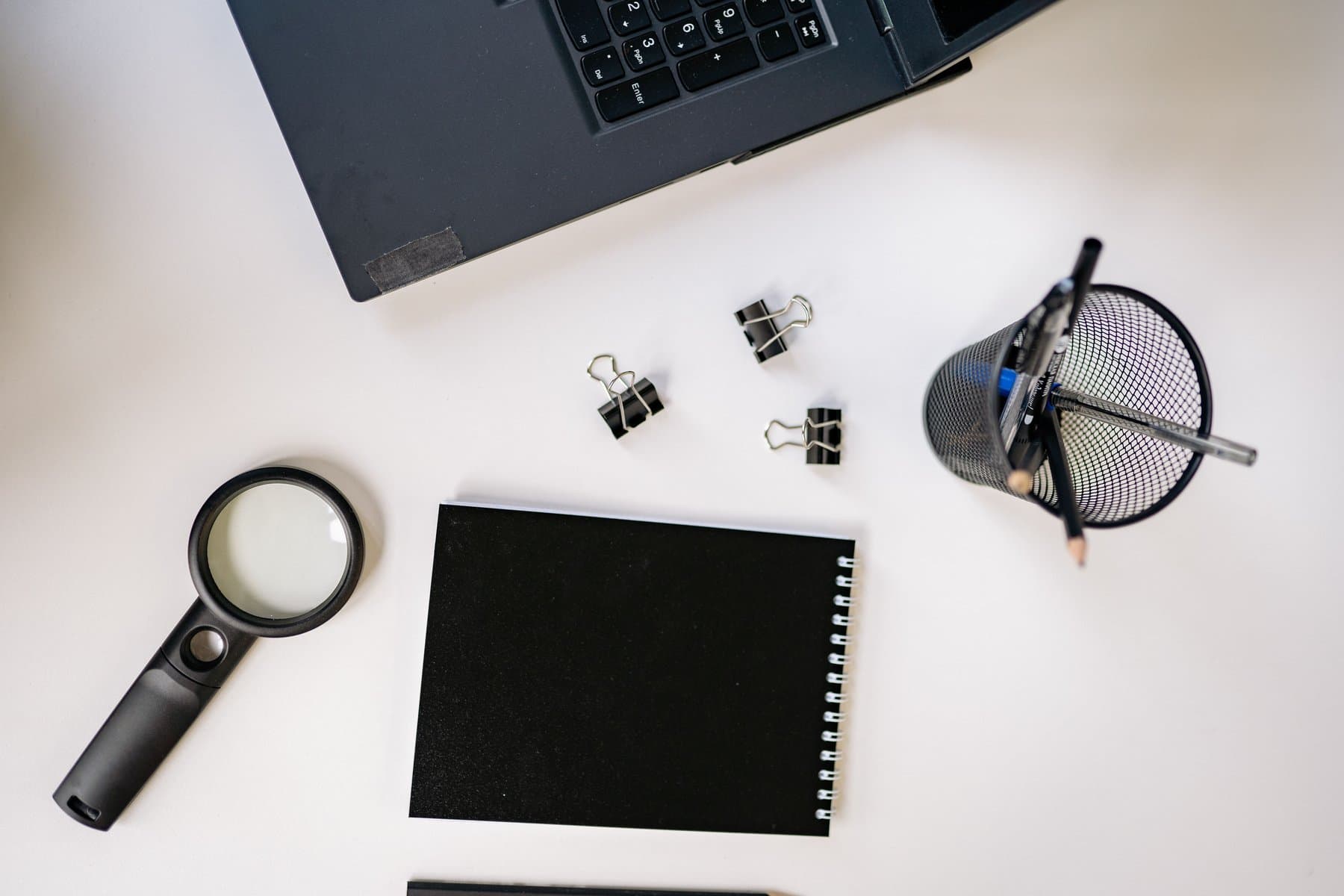 Magnifying glass, black notebook and desk supplies arranged on a desk seen from above next to a laptop