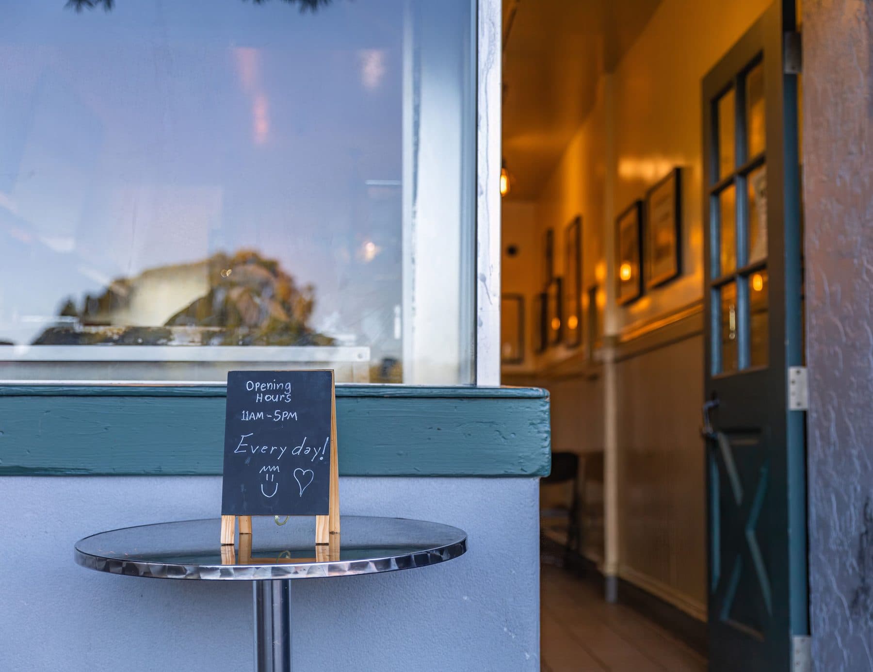 A small chalkboard reading 'Opening Hours, 11am-5pm, Every day' on a table outside a café, with the open entrance door in the background