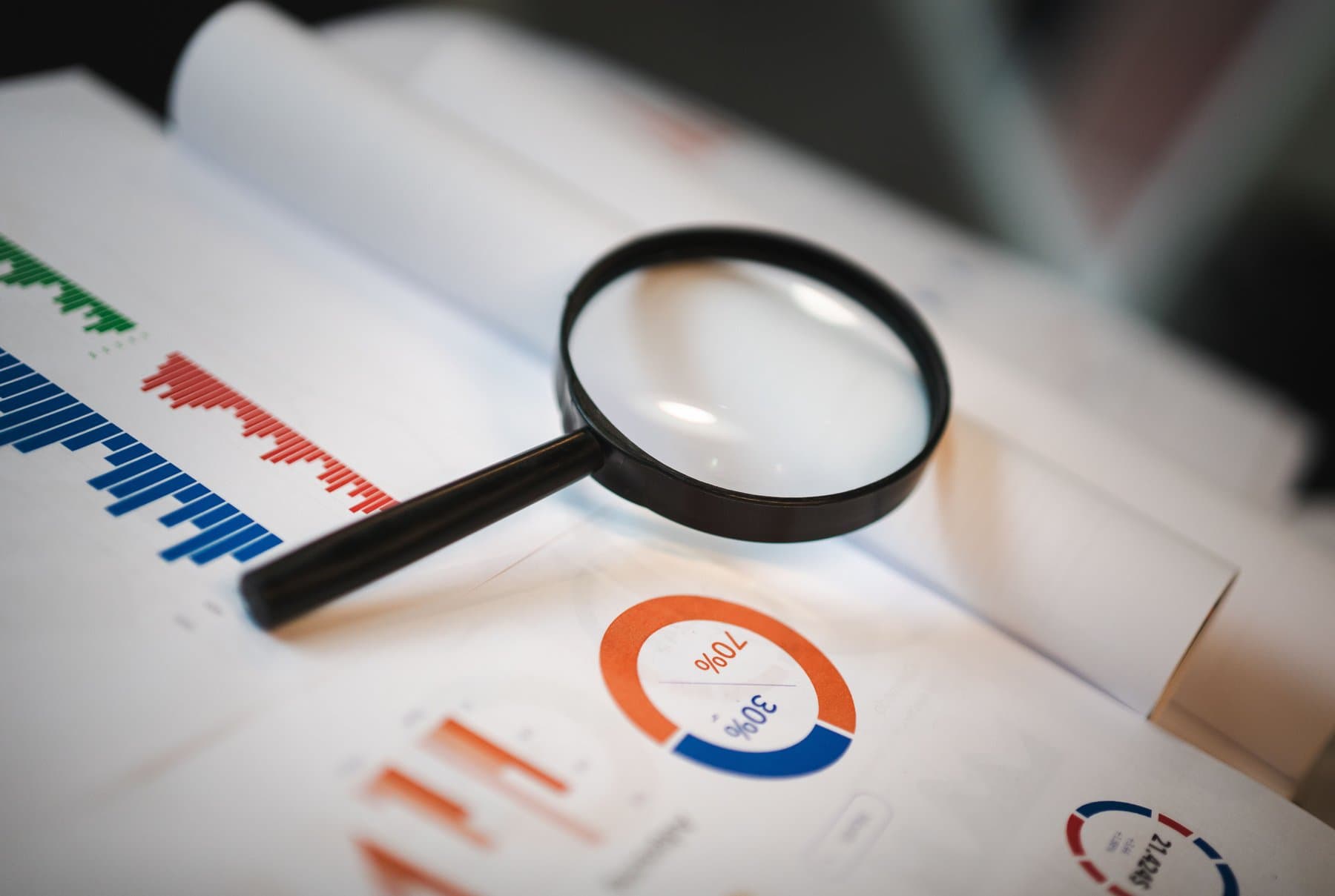 A magnifying glass resting on a printed sheet full of colourful bar charts and a pie chart with percentages