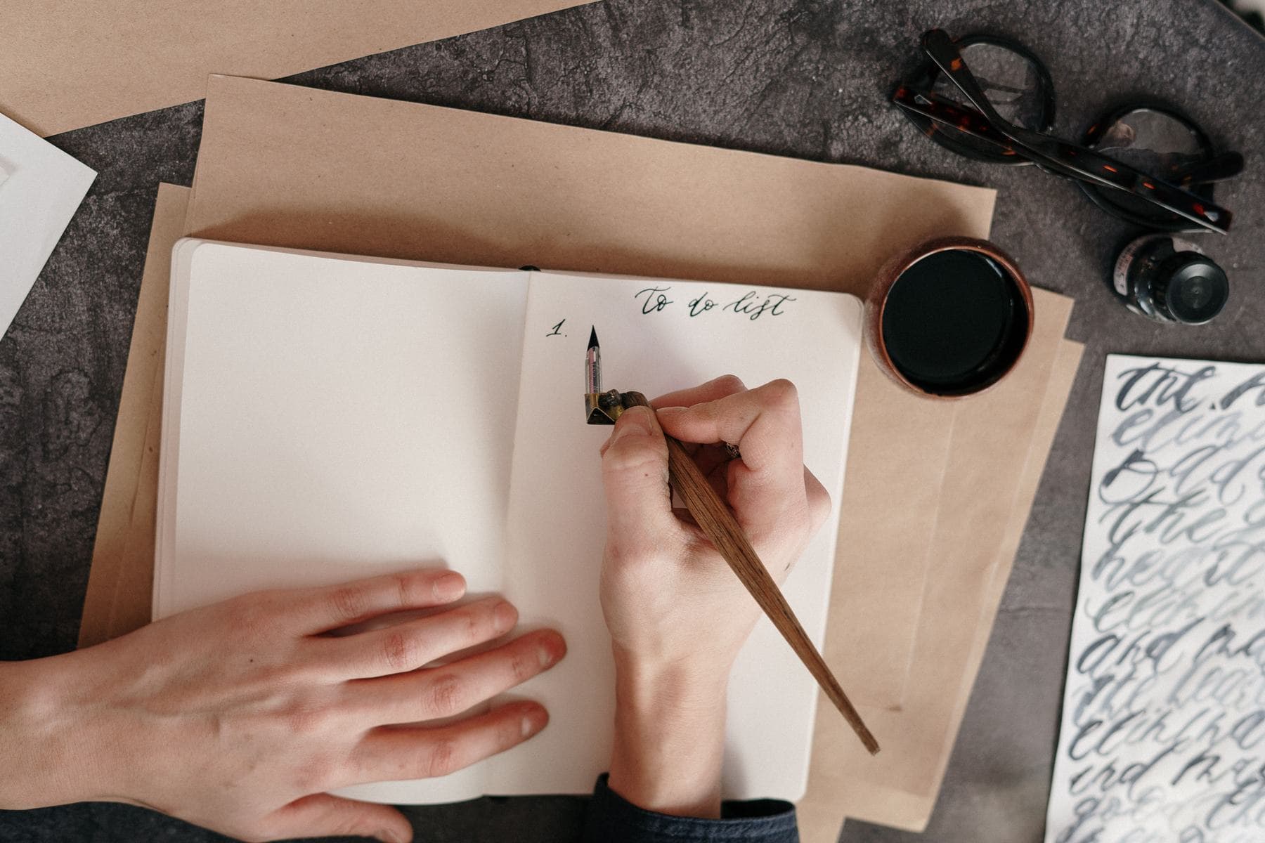 Top view of a hand writing a to-do list with a calligraphy pen and ink, on a desk with glasses nearby