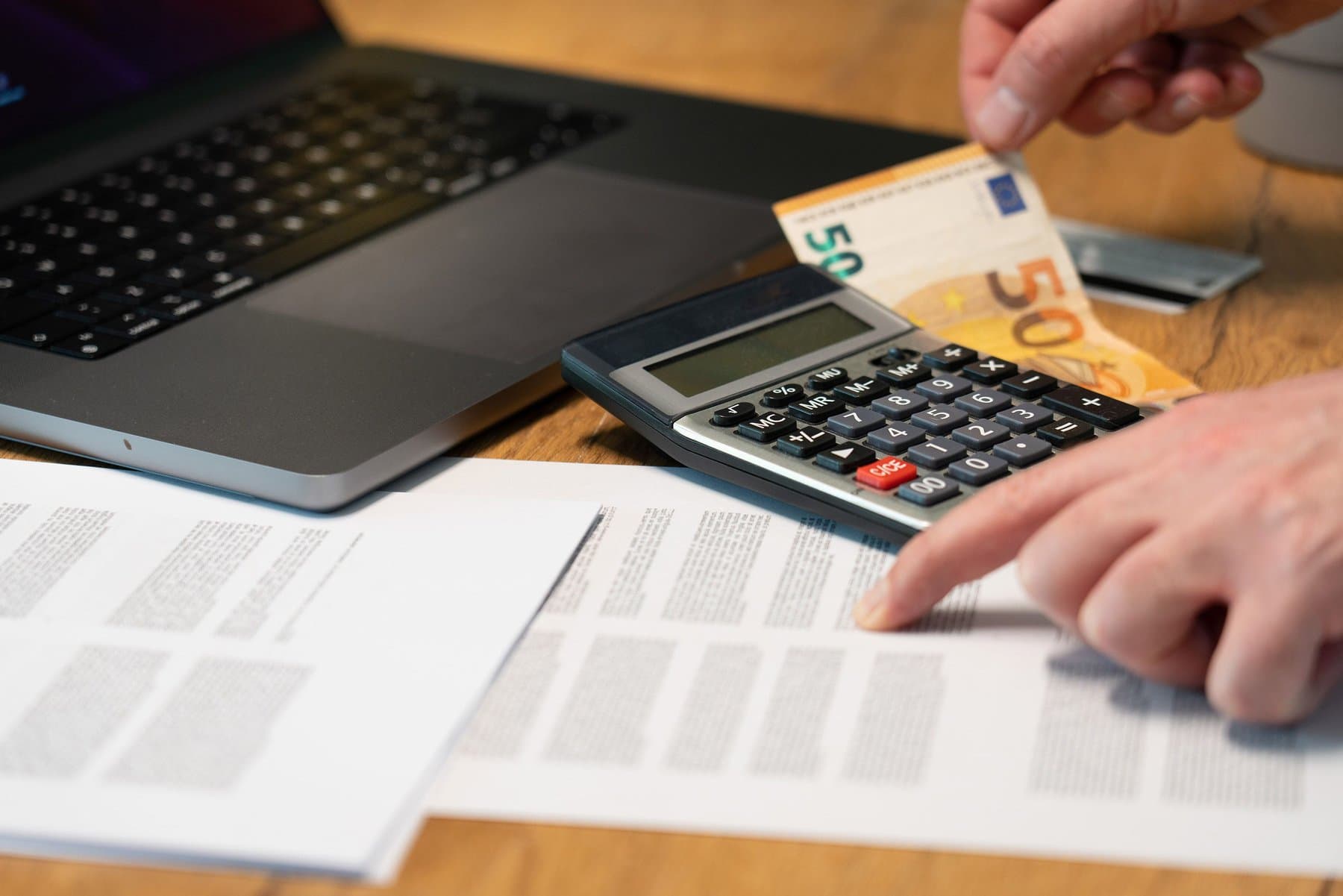 Una mano regge una banconota da 50 euro sopra una calcolatrice, accanto a un laptop chiuso e fogli stampati su una scrivania