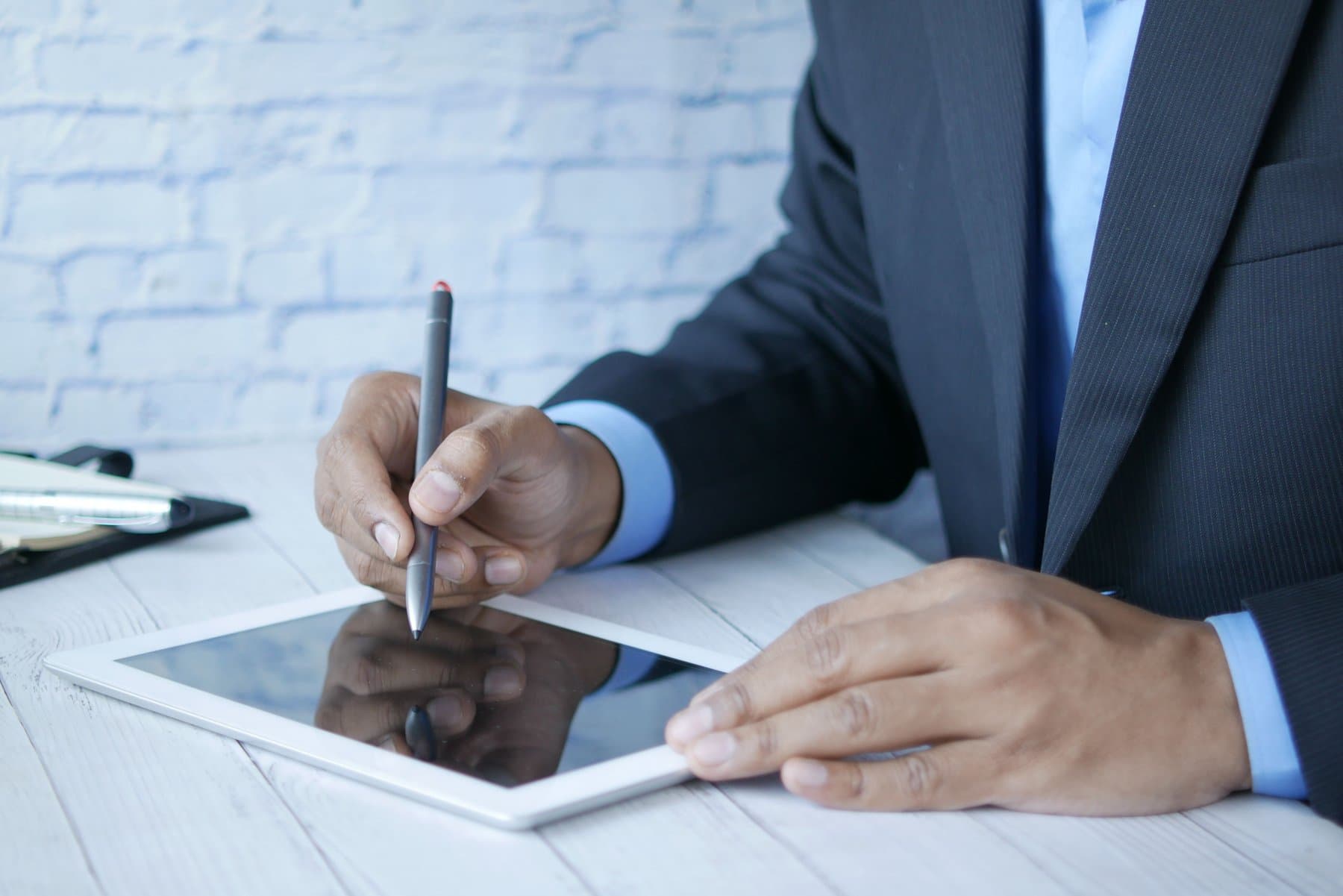 Close-up of a man in a suit holding a stylus over a white tablet, on a light wood desk