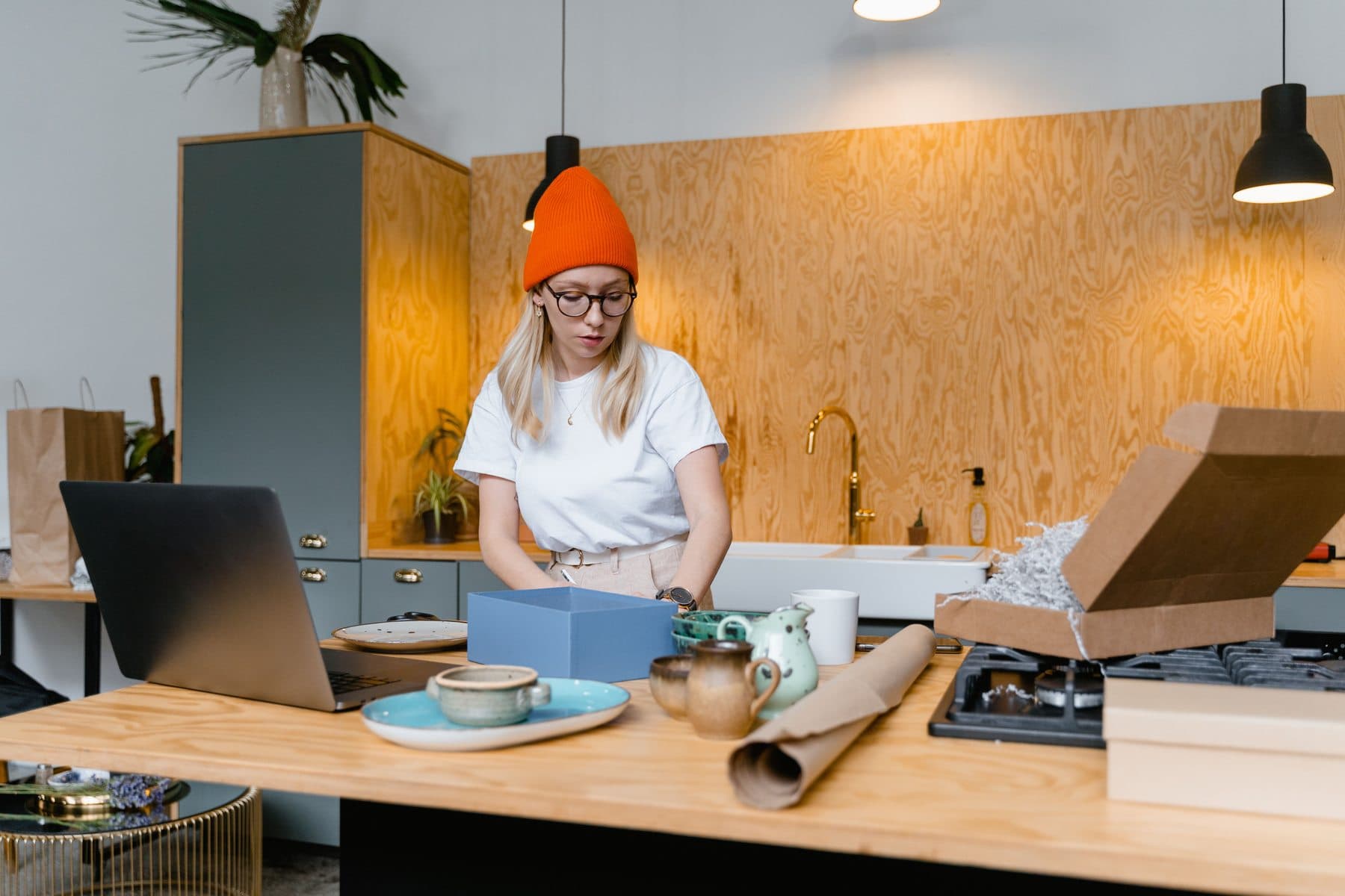 Una donna con un berretto arancione confeziona ceramiche fatte a mano su un bancone di legno, con laptop e scatole pronte per la spedizione