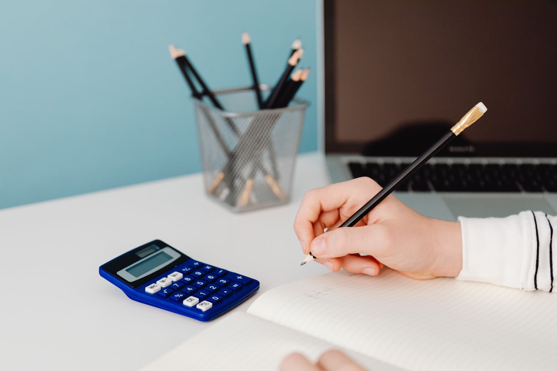 A hand writes with a pencil in a notebook, next to a blue calculator and a pencil cup, on a white desk