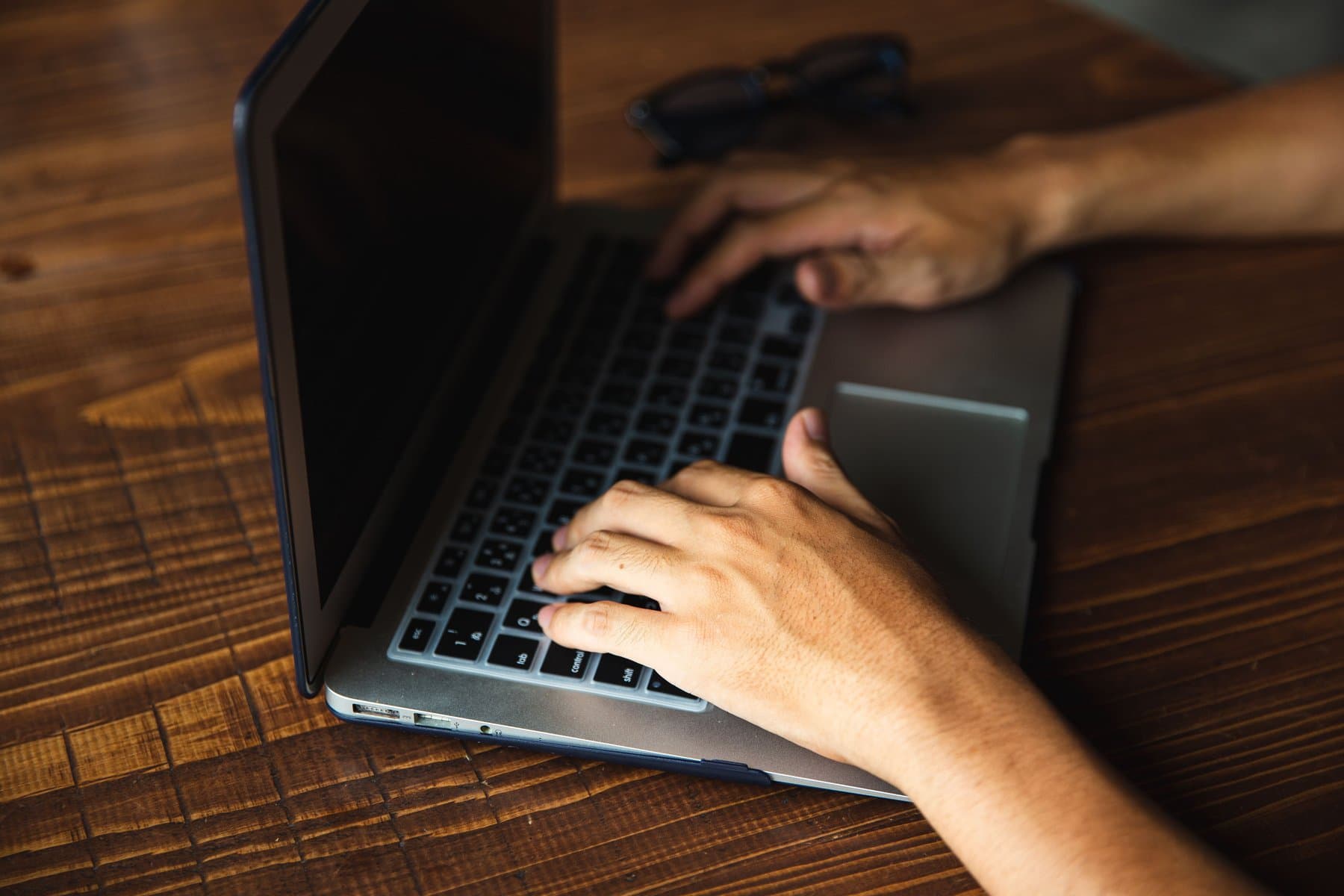 Due mani digitano su un laptop con lo schermo spento, appoggiato su un tavolo di legno, mentre un paio di occhiali resta sullo sfondo