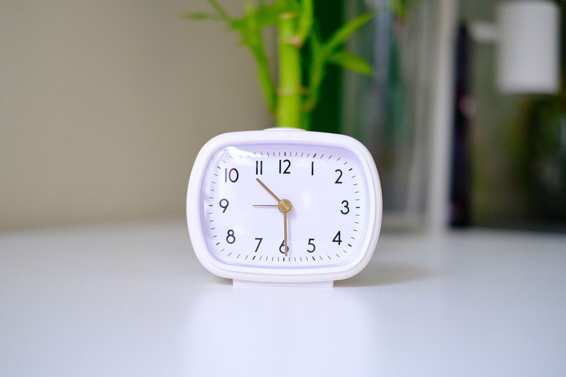 A minimalist white analogue clock on a clean desk, with a blurred plant in the background