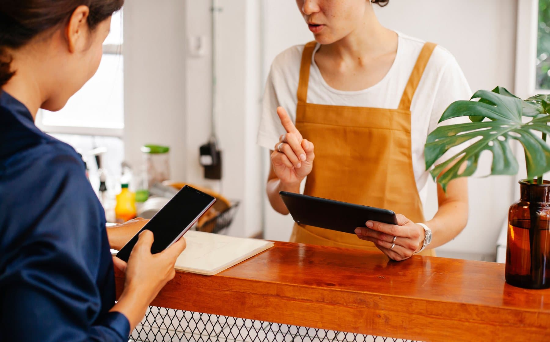 A shop assistant in an apron explains something to a customer at the counter, holding a tablet