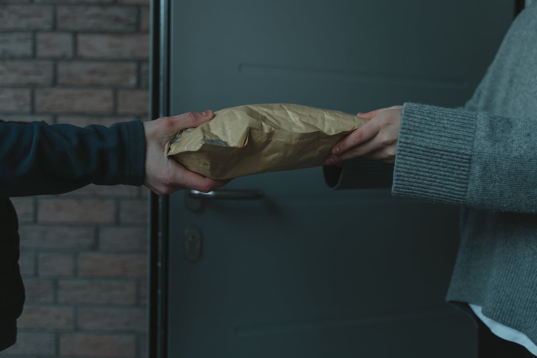 Primo piano di due mani che si scambiano un pacco chiuso in carta kraft sulla soglia di una casa