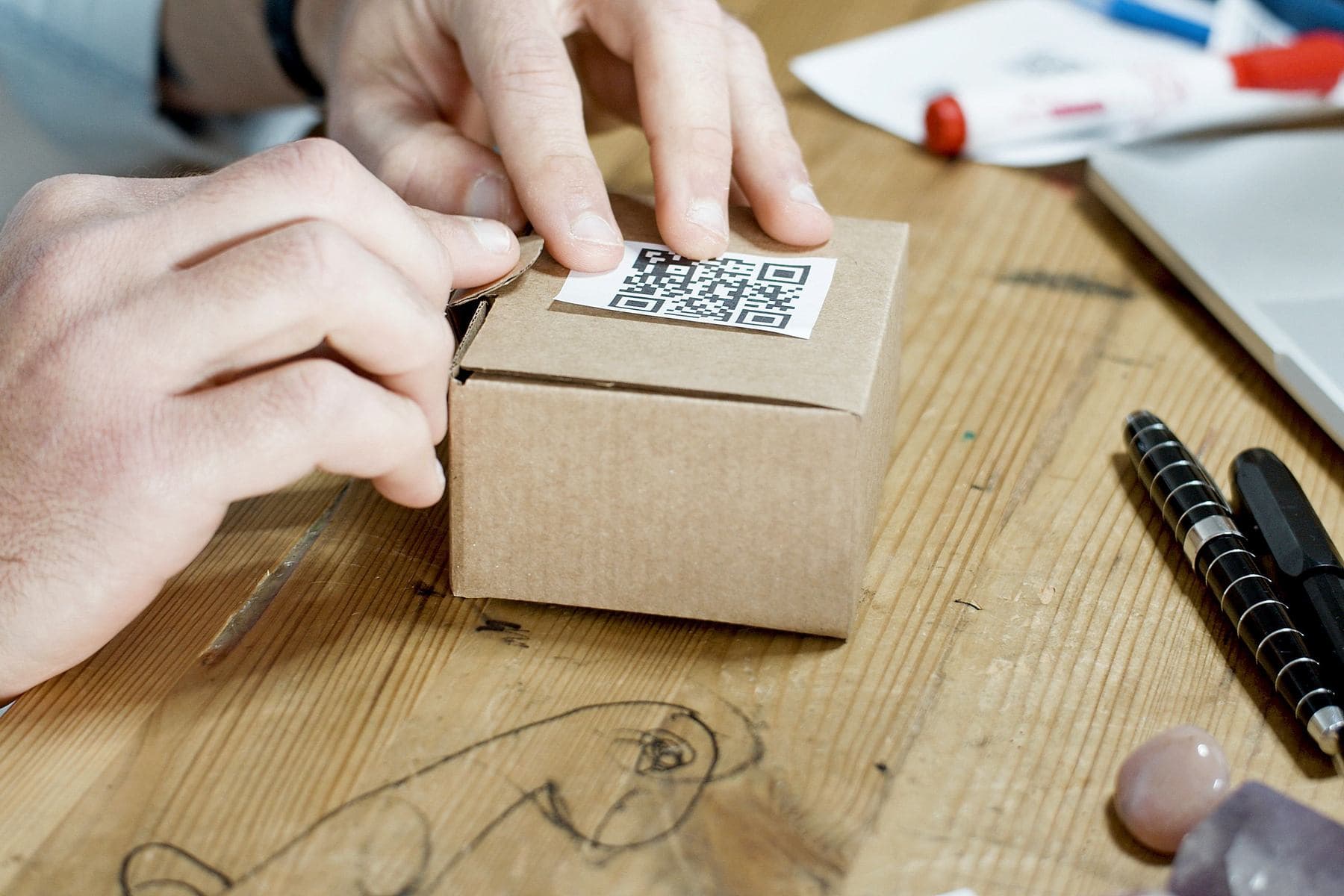 Mani che applicano un adesivo con codice QR sul coperchio di una piccola scatola di cartone, su una scrivania in legno