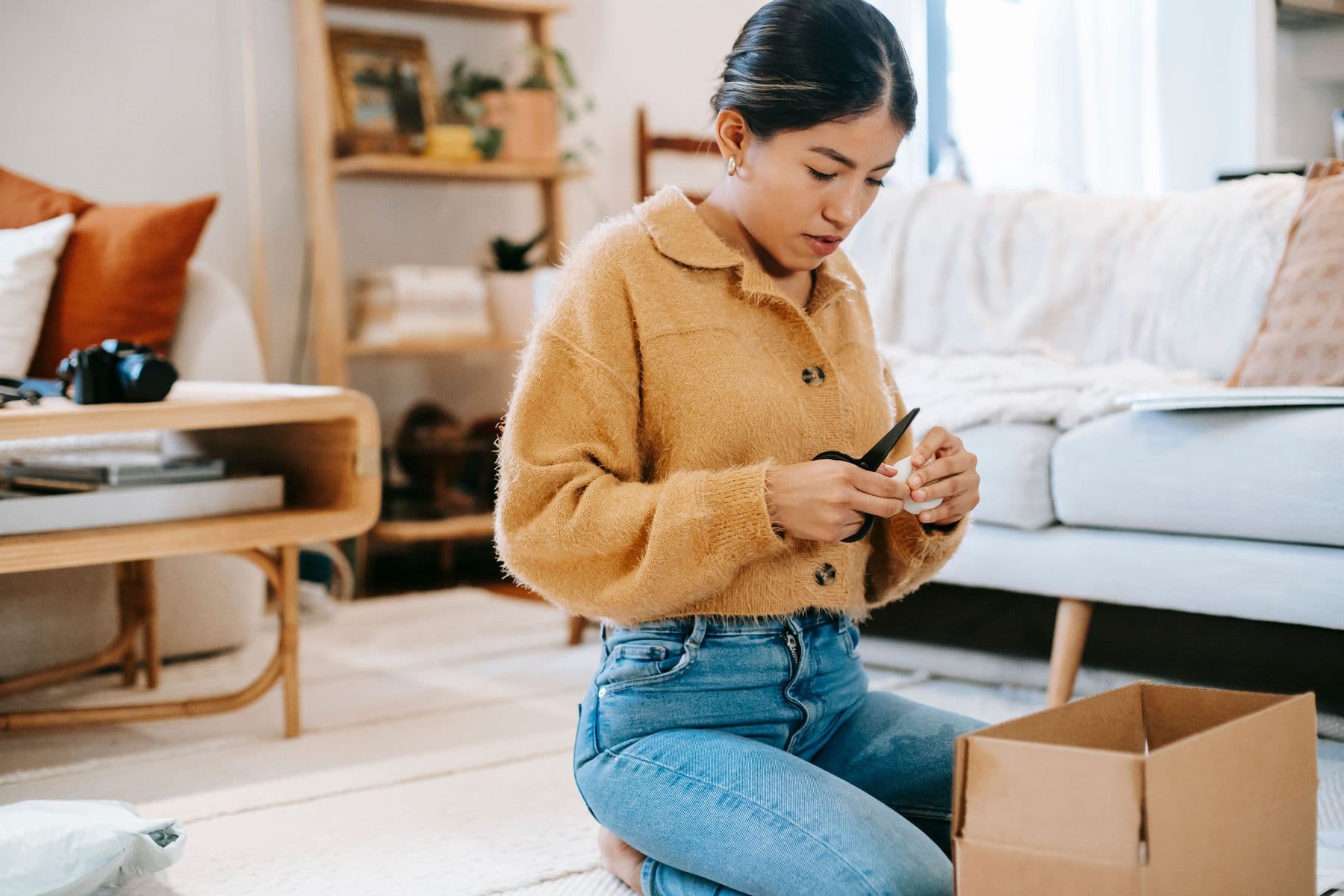 Young woman kneeling on a living room rug, leaning over an open cardboard box while using scissors