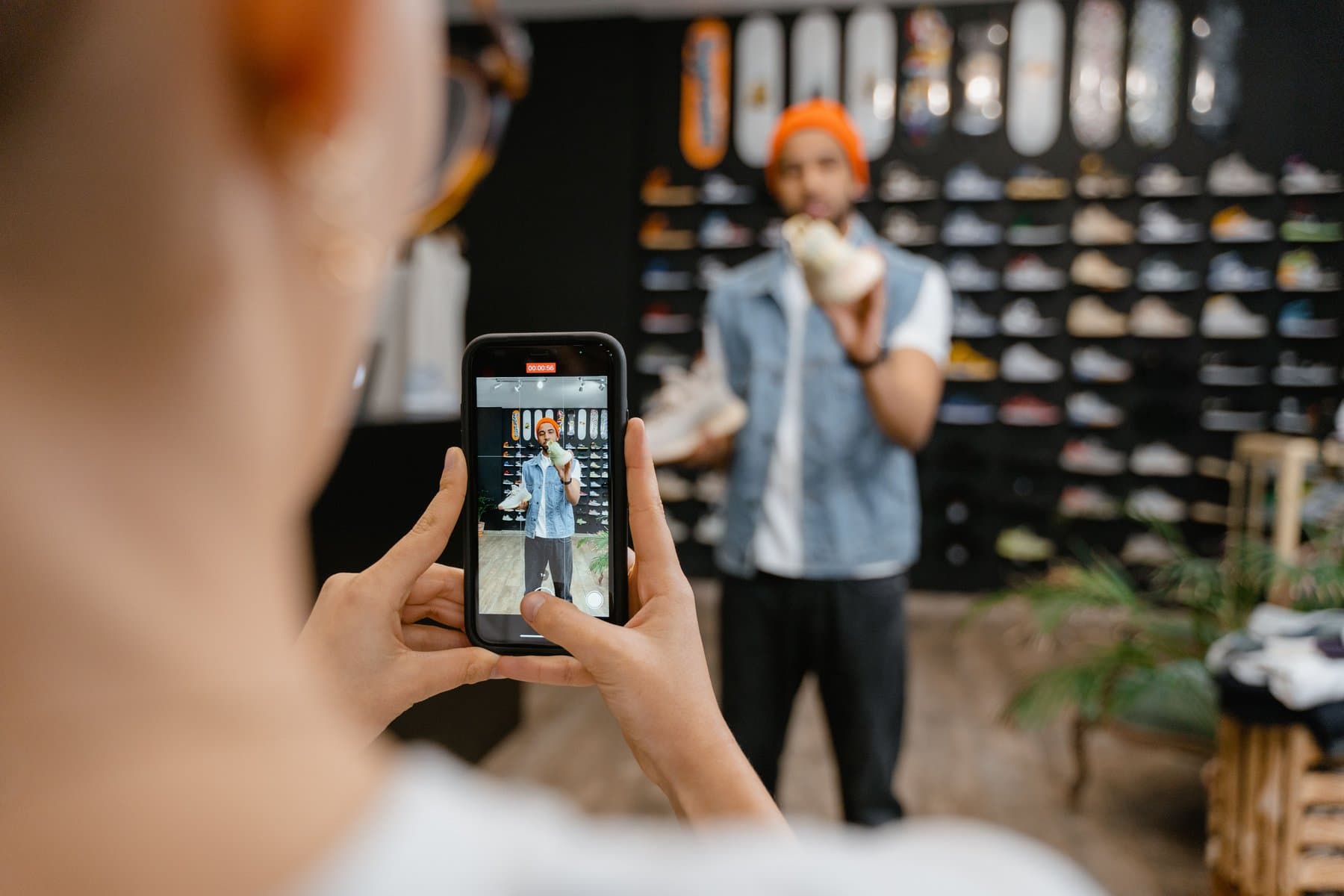 Una persona riprende con lo smartphone un'altra persona che mostra un paio di scarpe in un negozio, con il muro di scarpe esposte sullo sfondo
