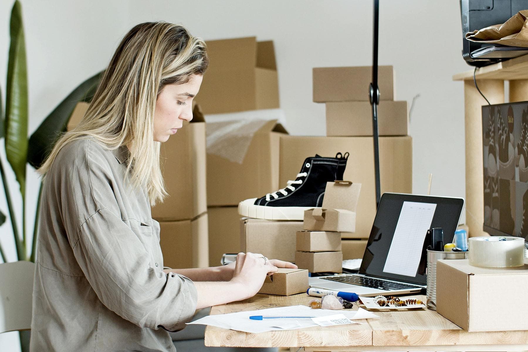 A woman packs a box at a work table, surrounded by parcels, with an open laptop in front of her
