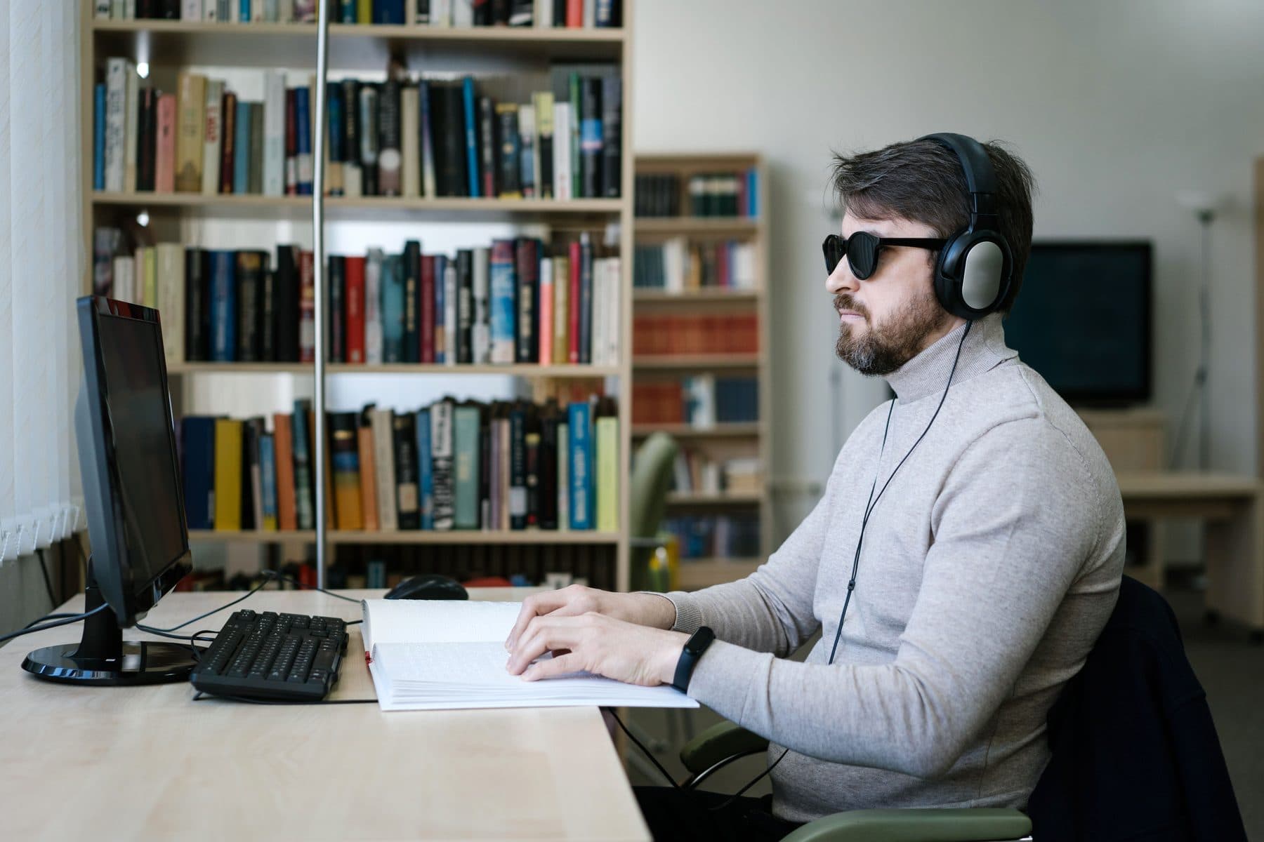 Un uomo con occhiali scuri e cuffie legge un testo in braille davanti al computer, in una biblioteca