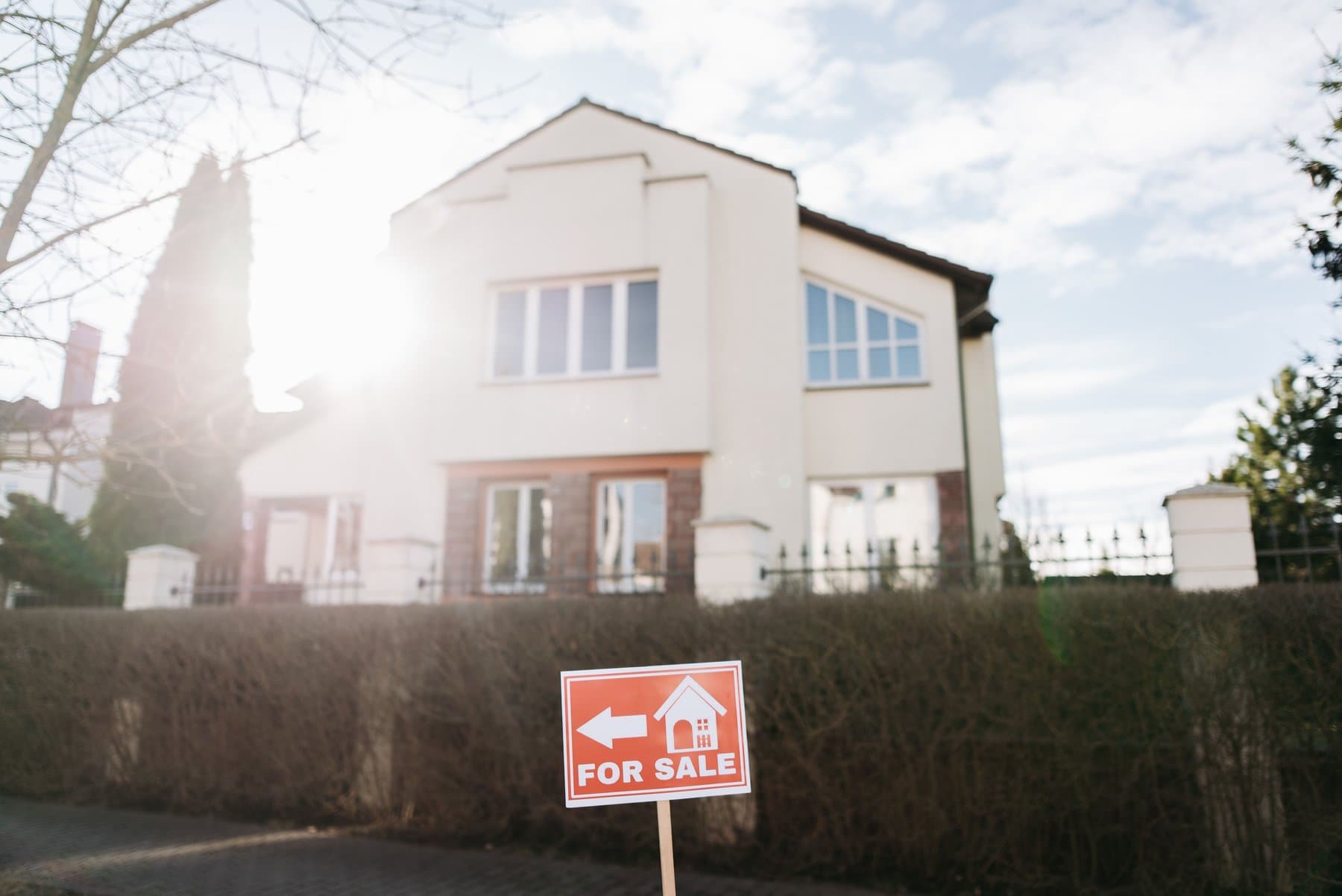 A 'for sale' sign in the foreground in front of a house with an iron fence and hedge, backlit at sunset