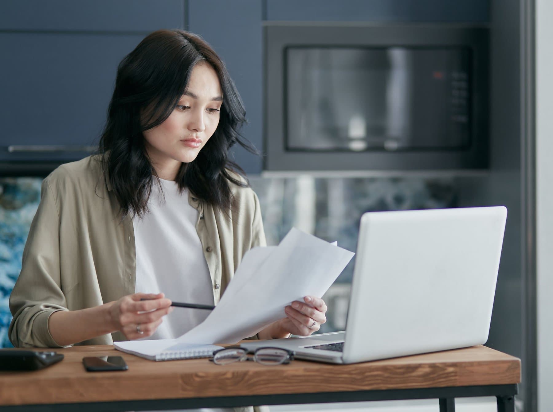 Donna concentrata che rivede dei documenti mentre lavora al laptop in un angolo cucina adibito a scrivania