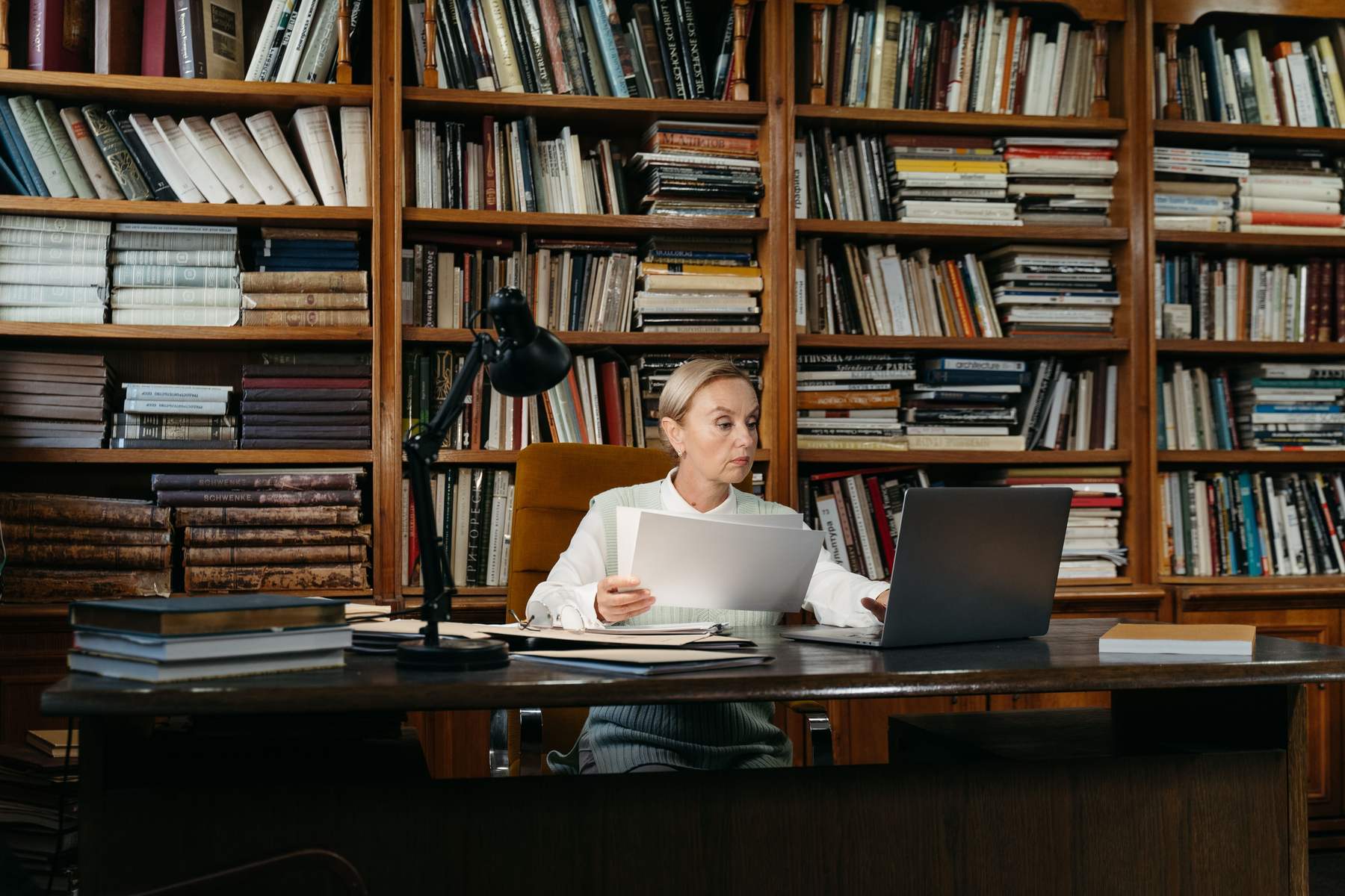 A professional reads documents in front of a laptop, sitting at a desk in an office lined with bookshelves