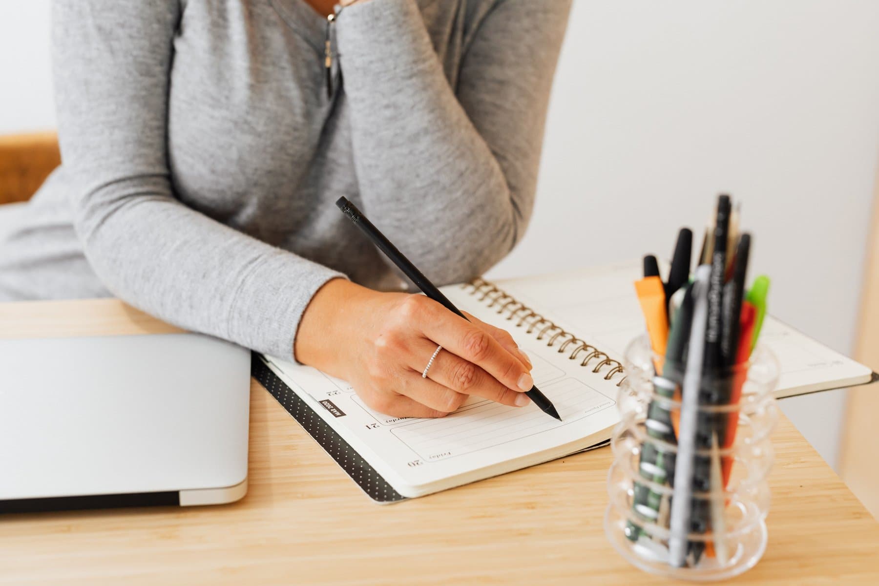 Una donna scrive a mano su un'agenda appoggiata su una scrivania di legno, con un laptop chiuso accanto e un portapenne pieno di pennarelli colorati sullo sfondo