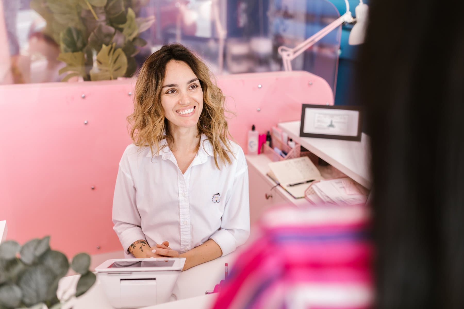 A receptionist smiles at a salon front desk in shades of pink, with a tablet and an open appointment book on the counter