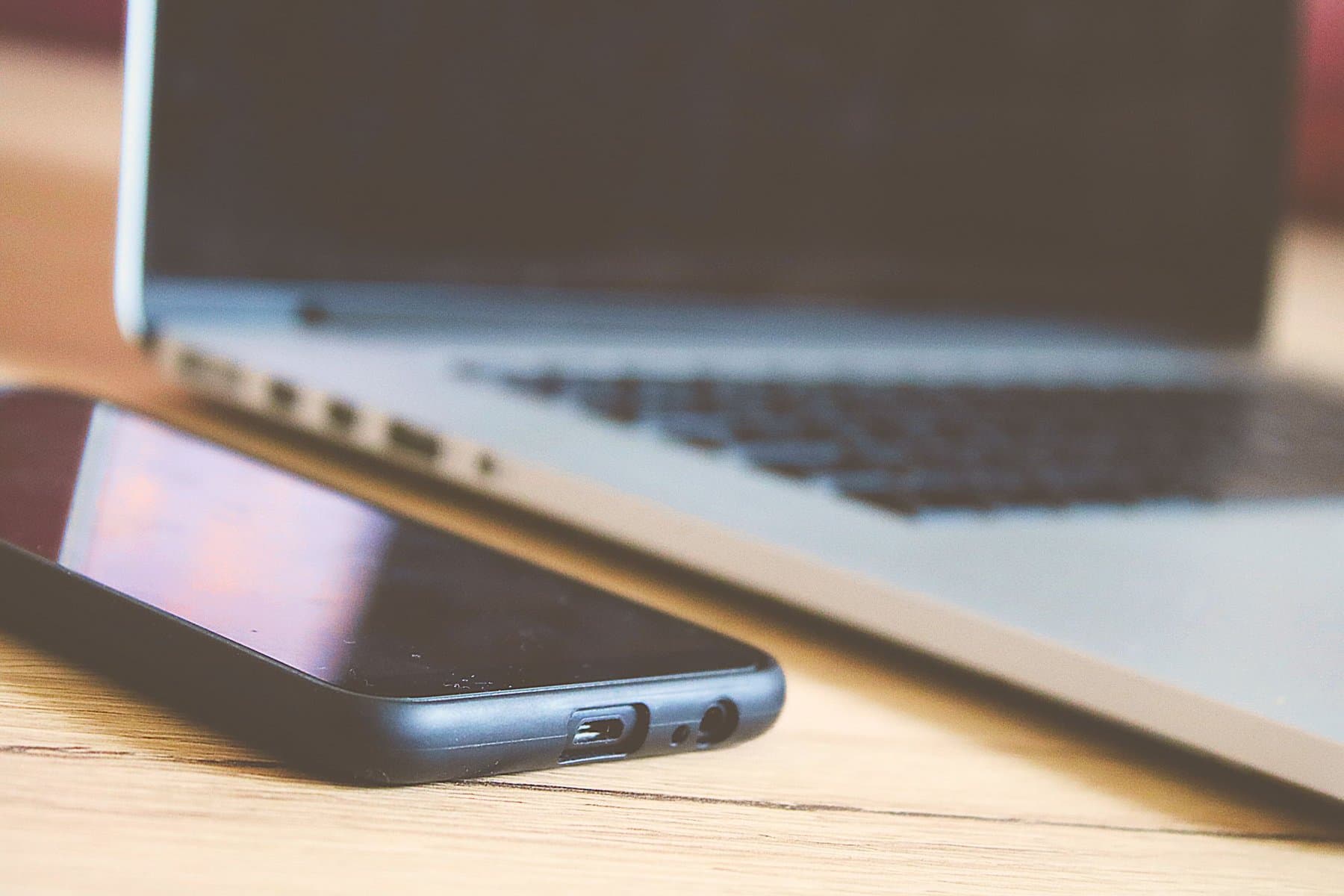 Close-up of a smartphone with its screen off resting on a wooden desk, with an open laptop blurred in the background