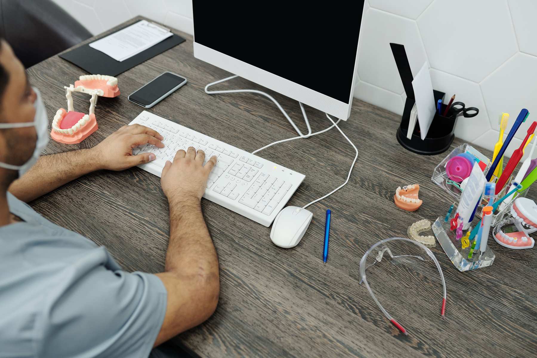 A dentist types on a computer keyboard, surrounded by dental models, colorful toothbrushes and tools on the practice desk