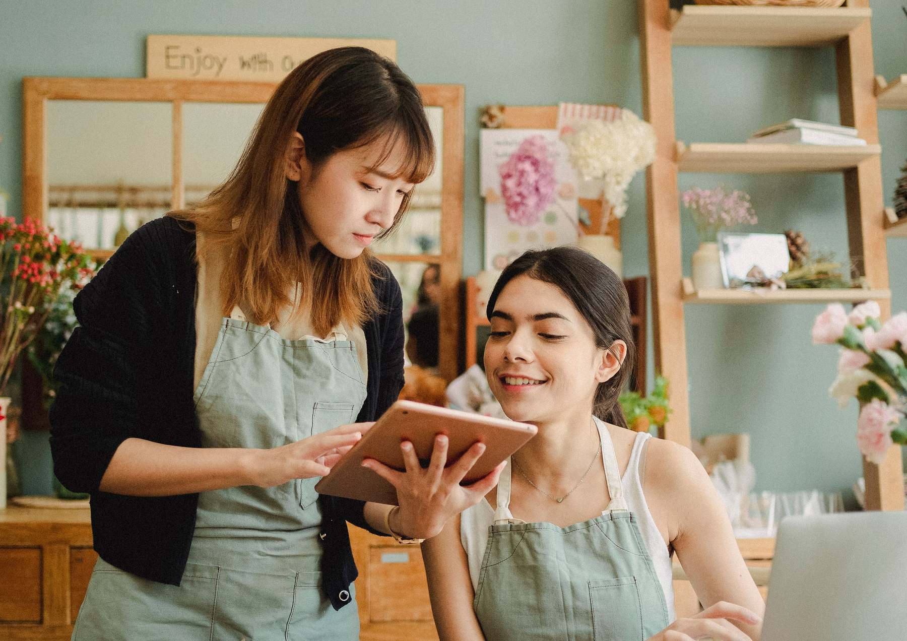 Two owners of a small flower shop look together at a tablet behind the counter, surrounded by flowers and wooden shelves