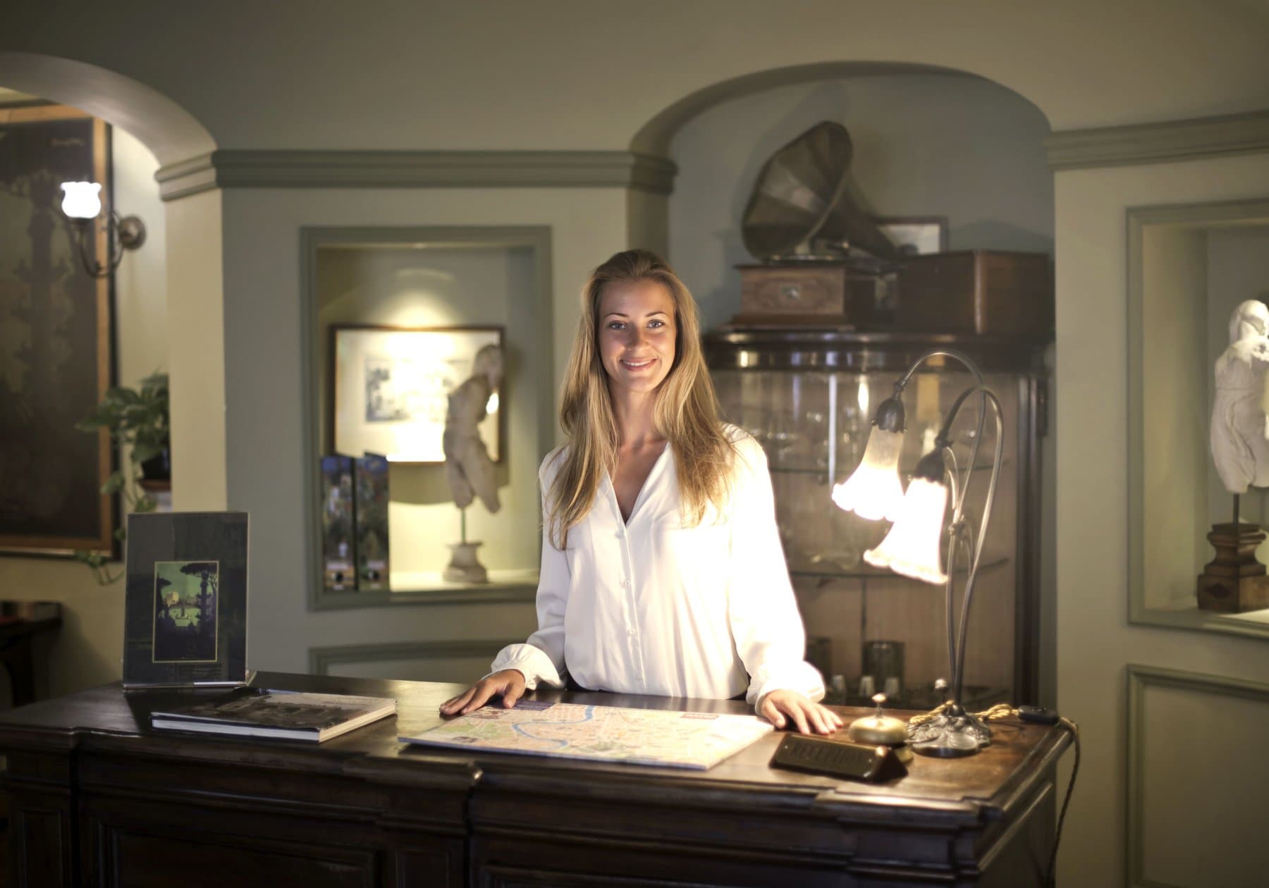A receptionist smiles behind the wooden desk of a period hotel, with a city map open on the counter