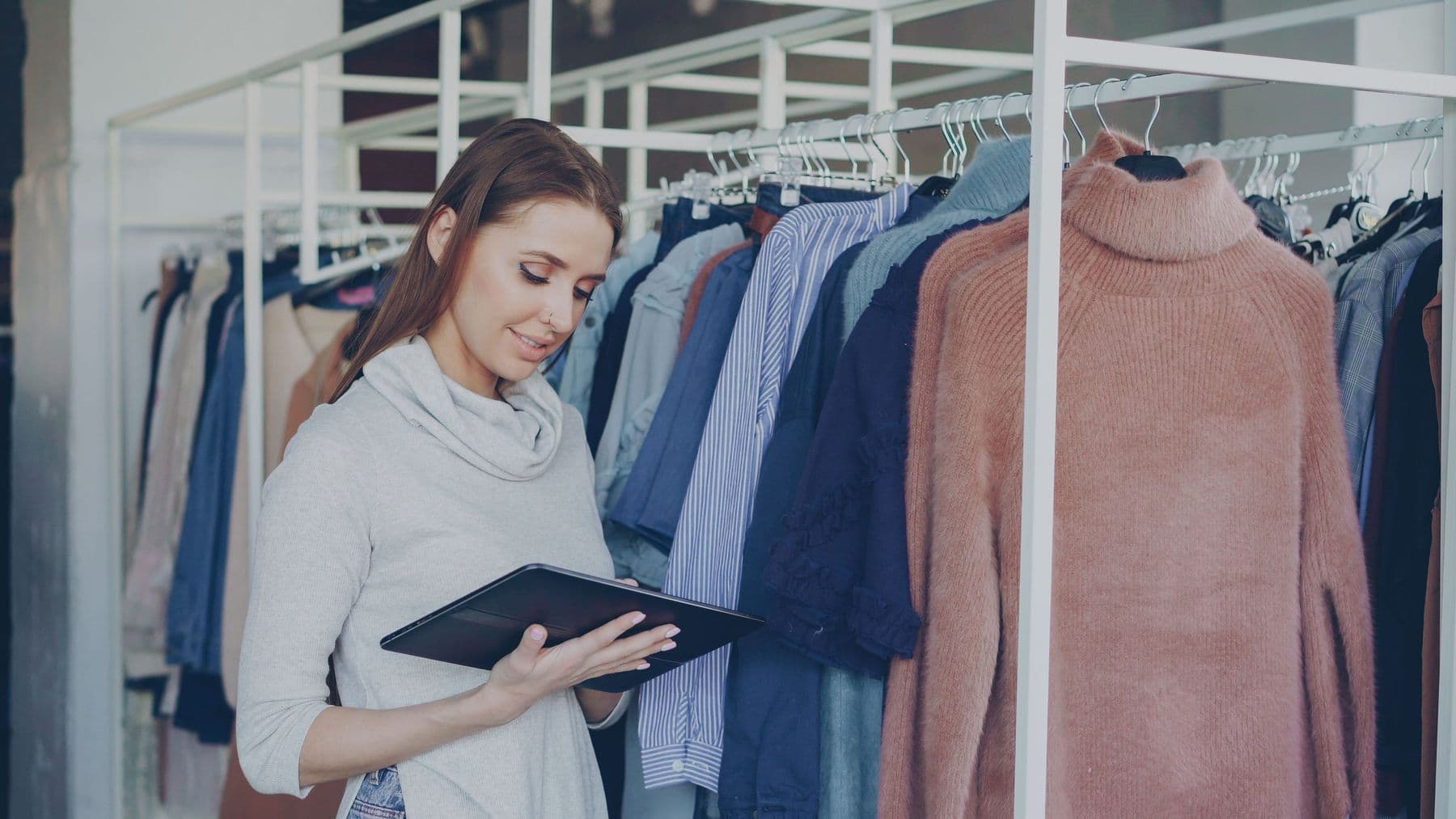 Shop assistant in a clothing store checking inventory on a tablet, in front of two clothing racks