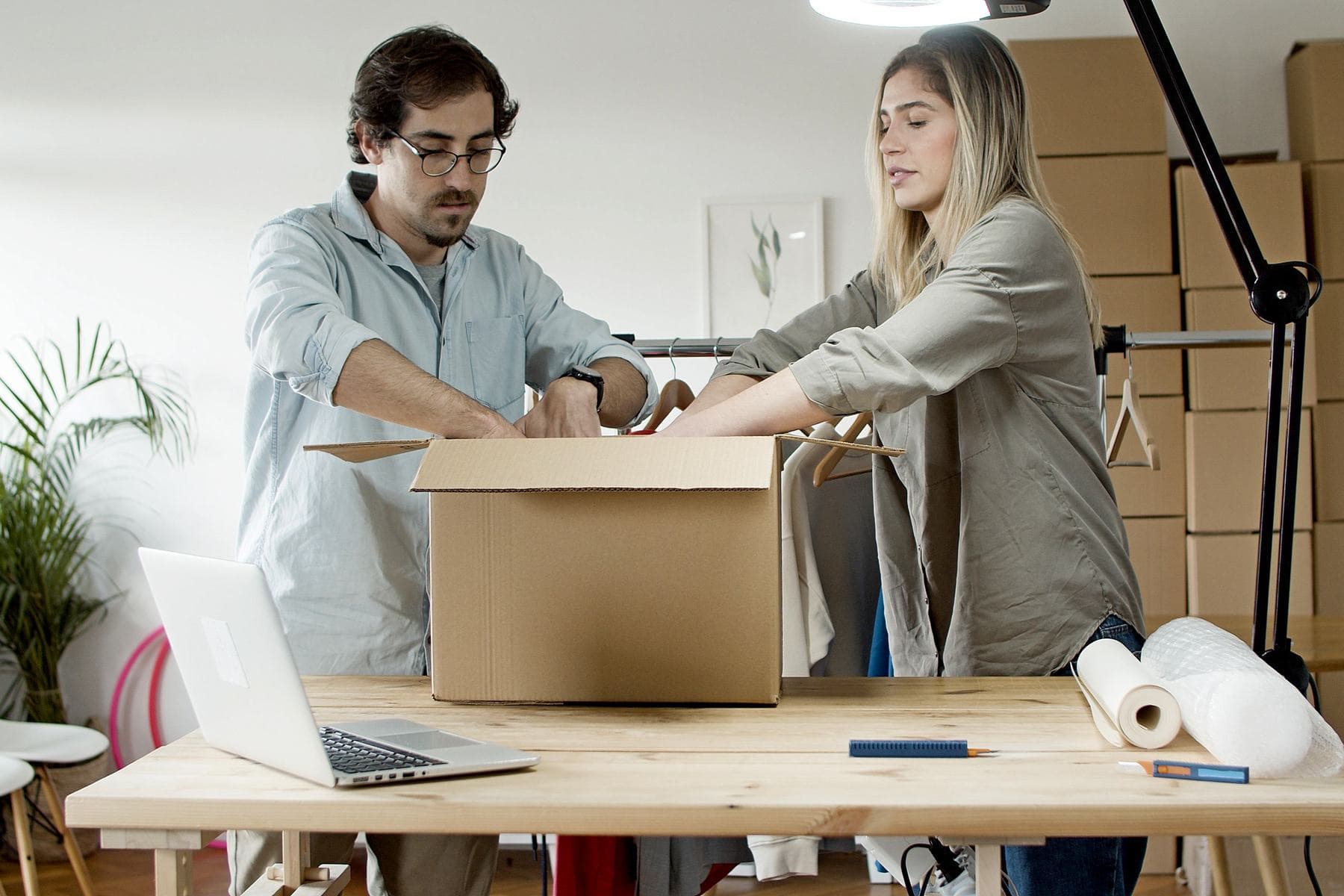 A man and a woman pack an order into a cardboard box together, at a work table with a laptop open and more boxes stacked in the background